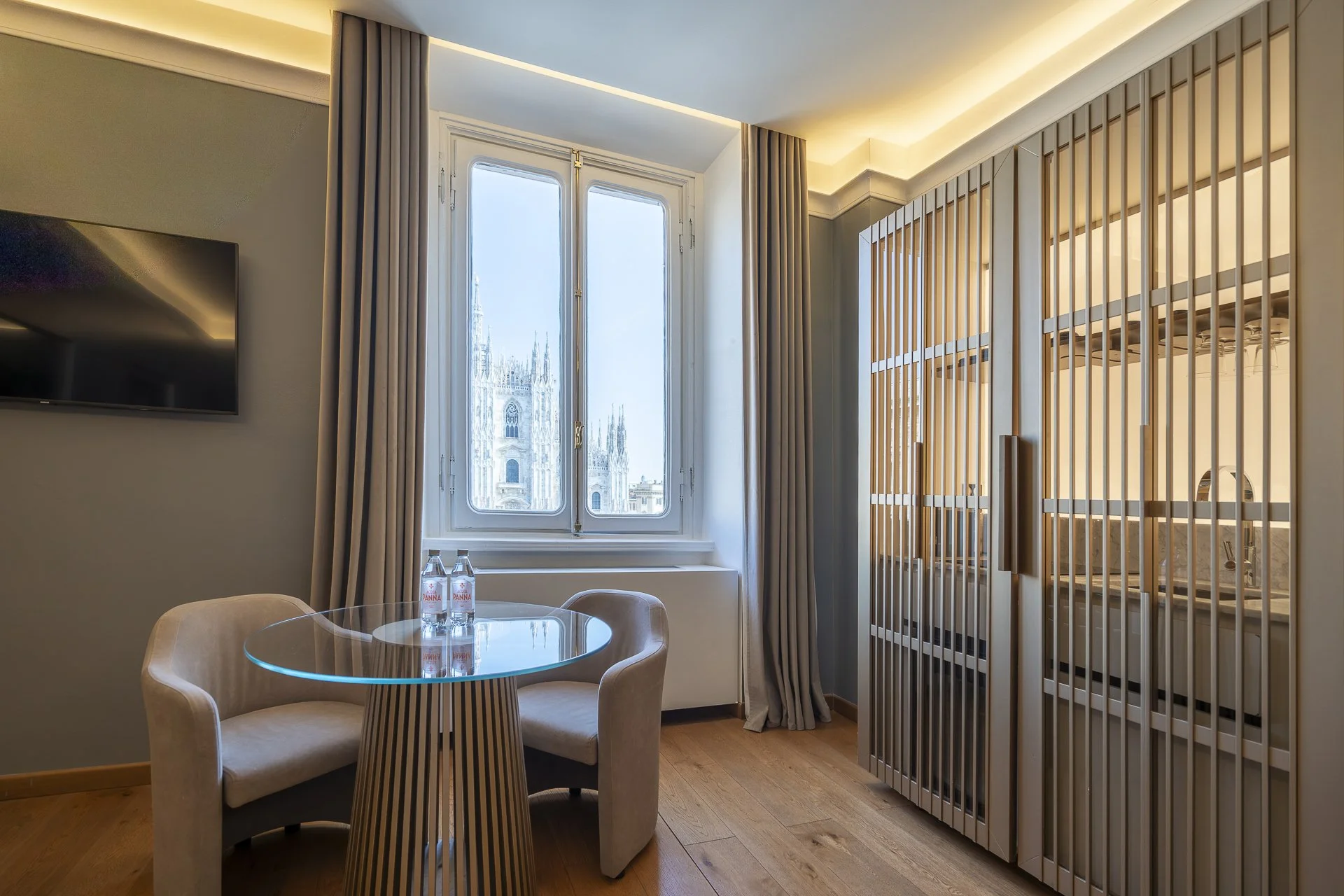 Interno di una stanza con finestra che mostra il duomo di Milano, con un tavolino di vetro e due sedie, e vaschette di acqua sul tavolino.