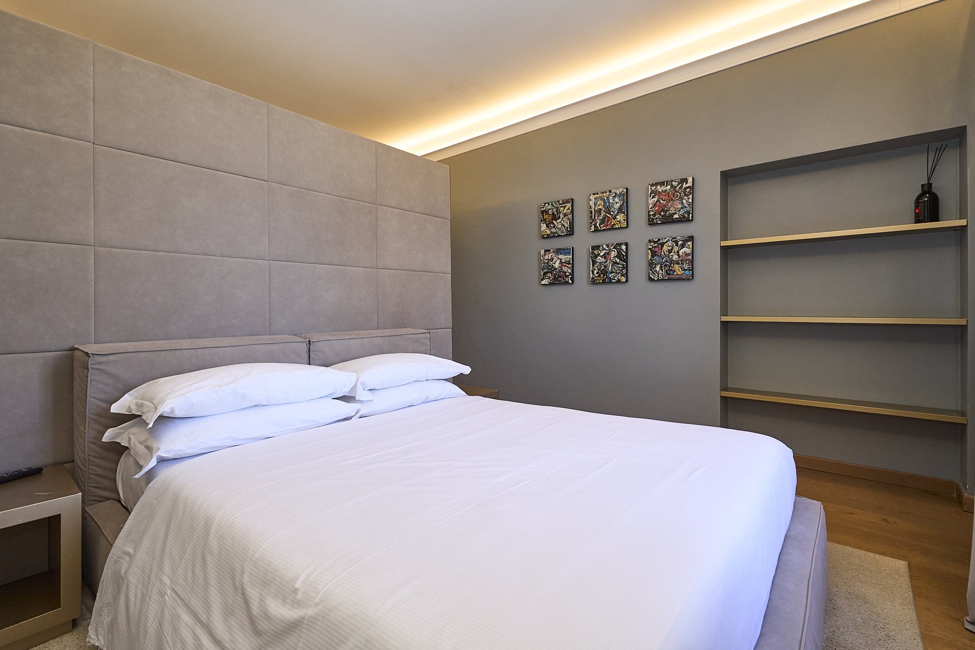 Camera da letto moderna con letto con biancheria bianca, parete con pannelli imbottiti, collage di cinque quadri astratti e scaffale vuoto con una decorazione nera.