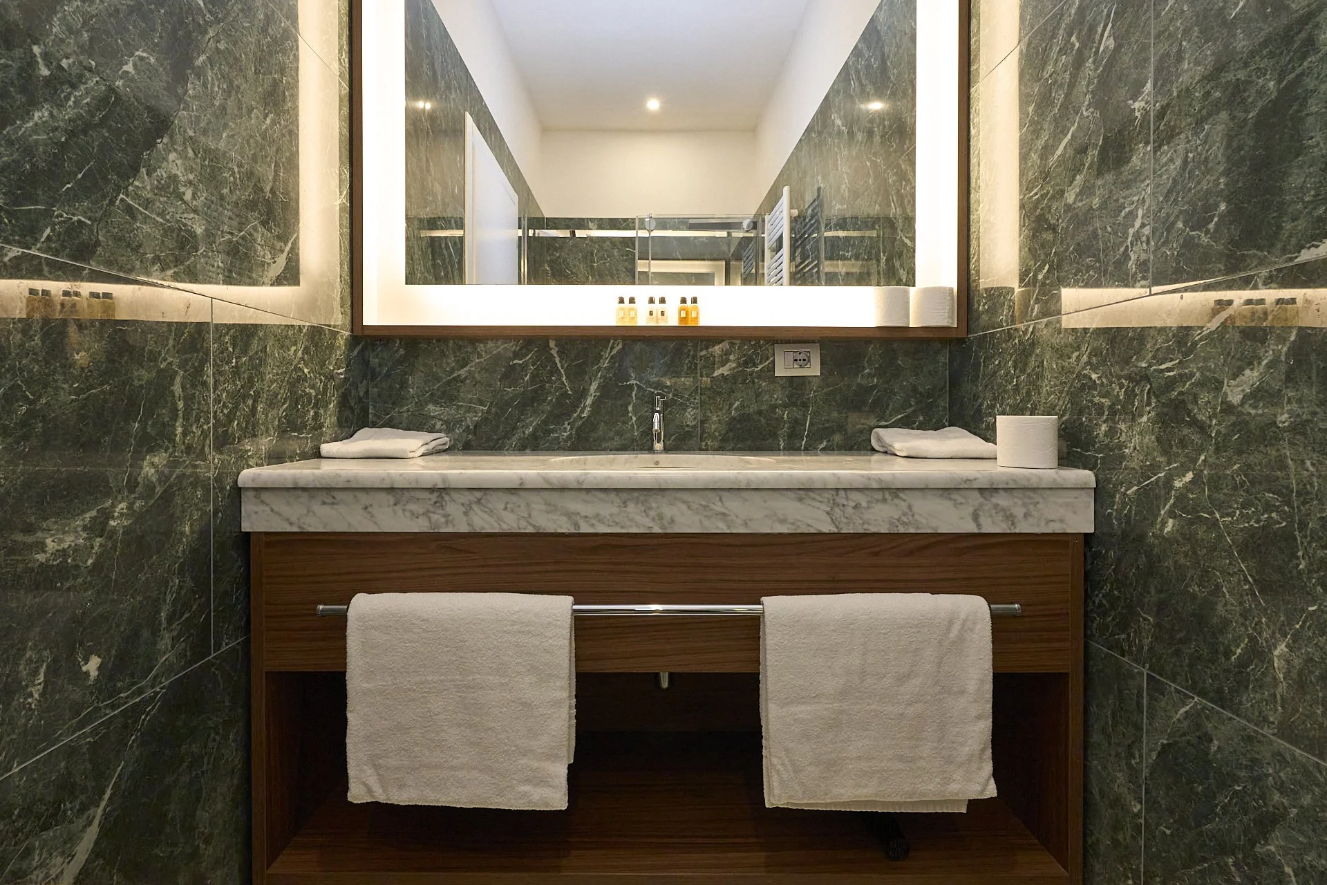 Lavabo con teli bianchi, specchio con luci e pareti in marmo verde scuro