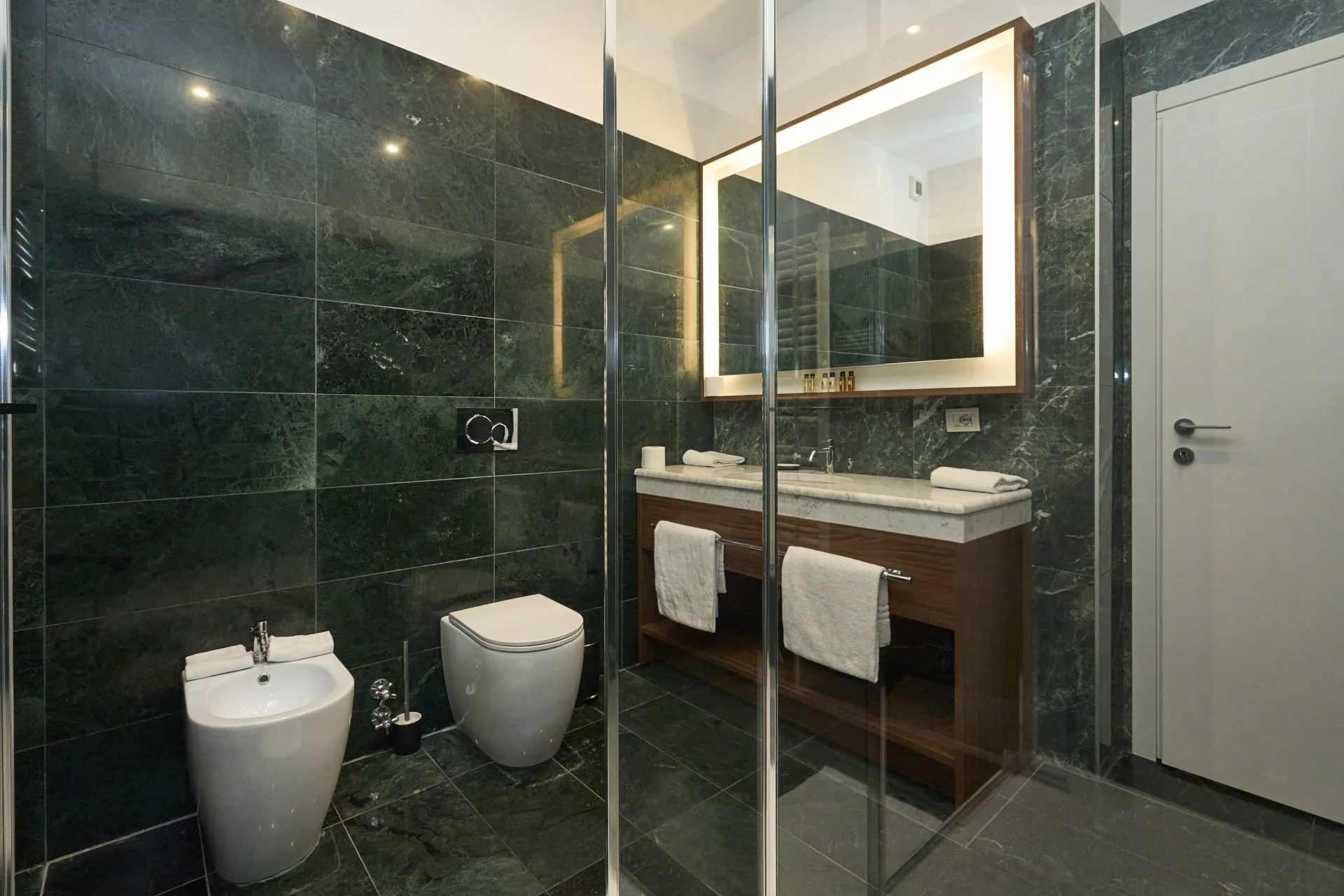Bagno moderno con pareti e pavimenti in marmo verde, lavabo con specchio retroilluminato, sanitari bianchi, e porta di accesso a destra.