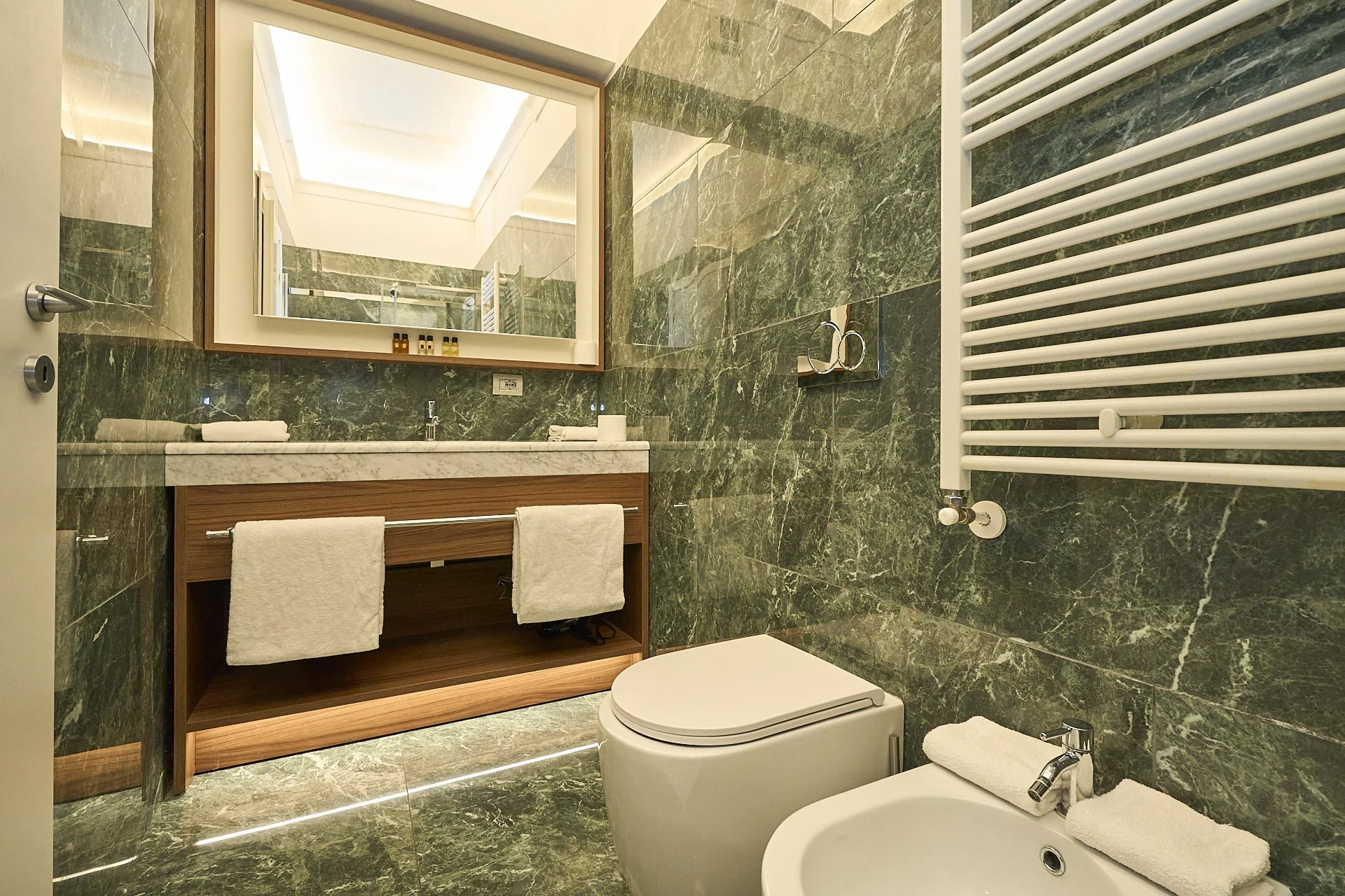 Bagno con pareti e pavimenti in marmo verde, grande specchio sopra il lavabo con ripiano in marmo, asciugamani bianchi appesi, riscaldamento a radiatore bianco, tazza del WC e bidet con asciugamani sopra, piccole bottiglie di prodotti da bagno sul ri