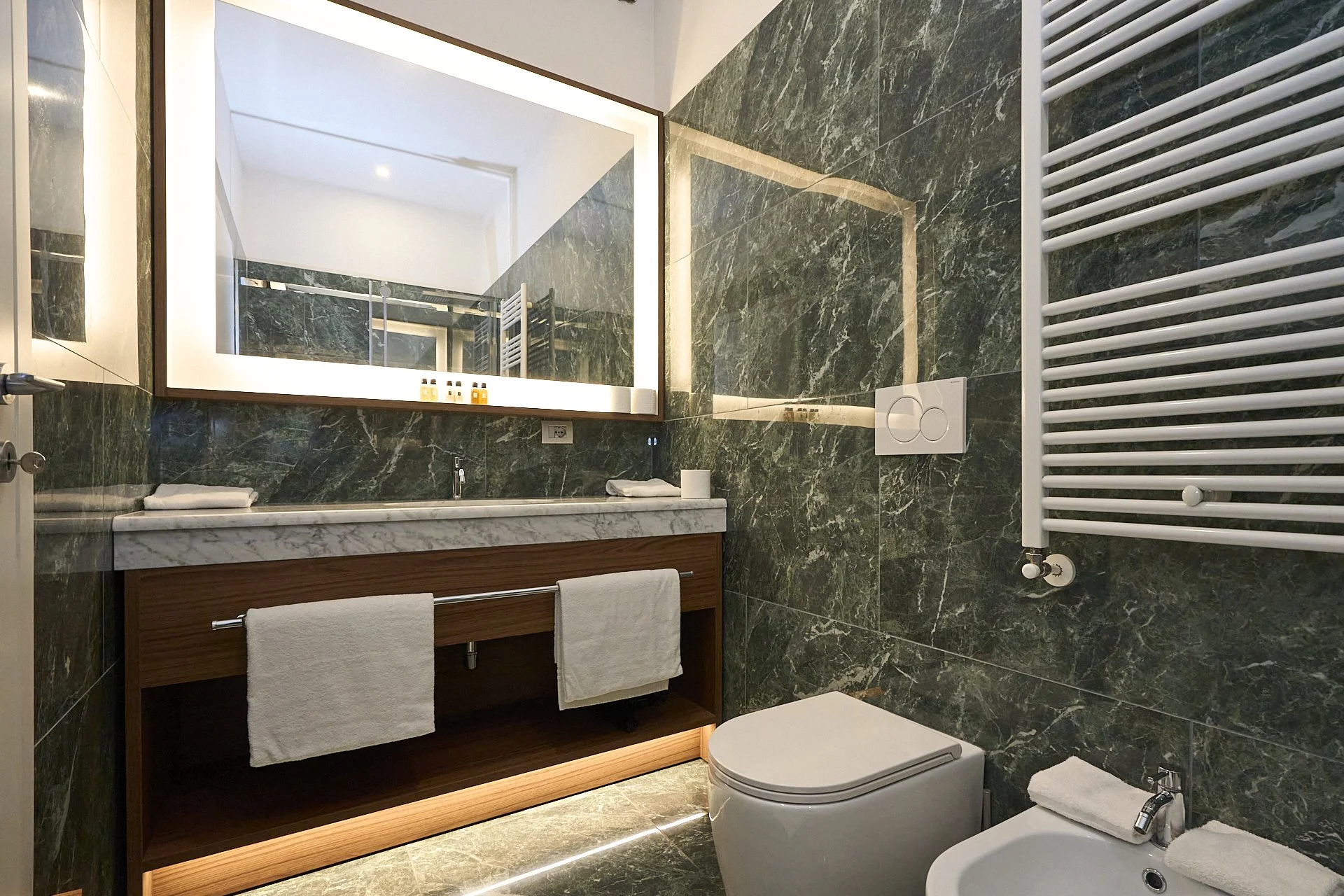 Bagno moderno con pareti e pavimento in marmo verde scuro, grande specchio con illuminazione retroilluminata, lavabo su mobile in legno con asciugamani, water e bidet, radiatore a caldo in alto a destra.