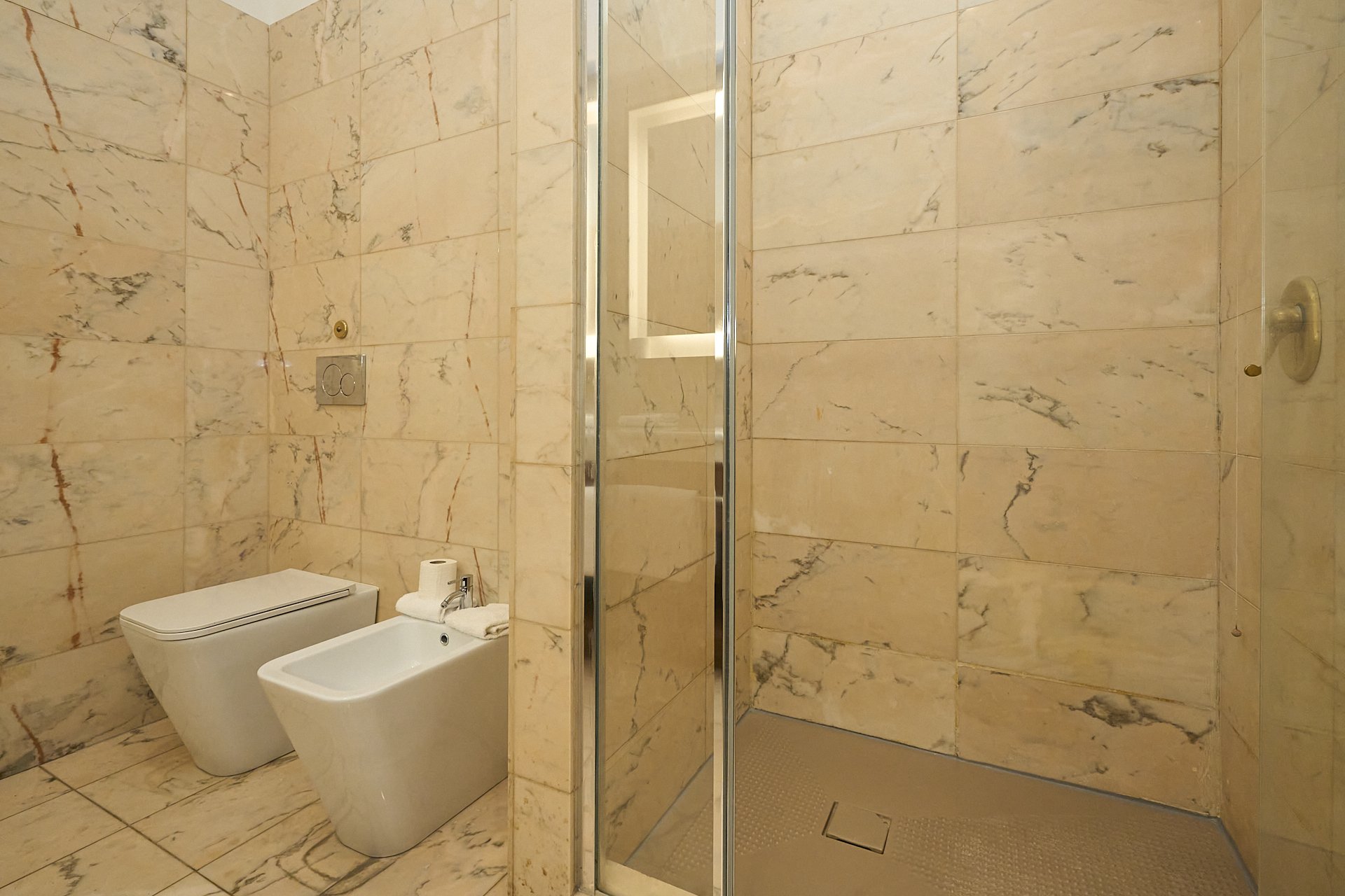 Bagno moderno con pareti e pavimenti in marmo chiaro, inclusi water e bidet, area doccia con porta in vetro e alte pareti in marmo.