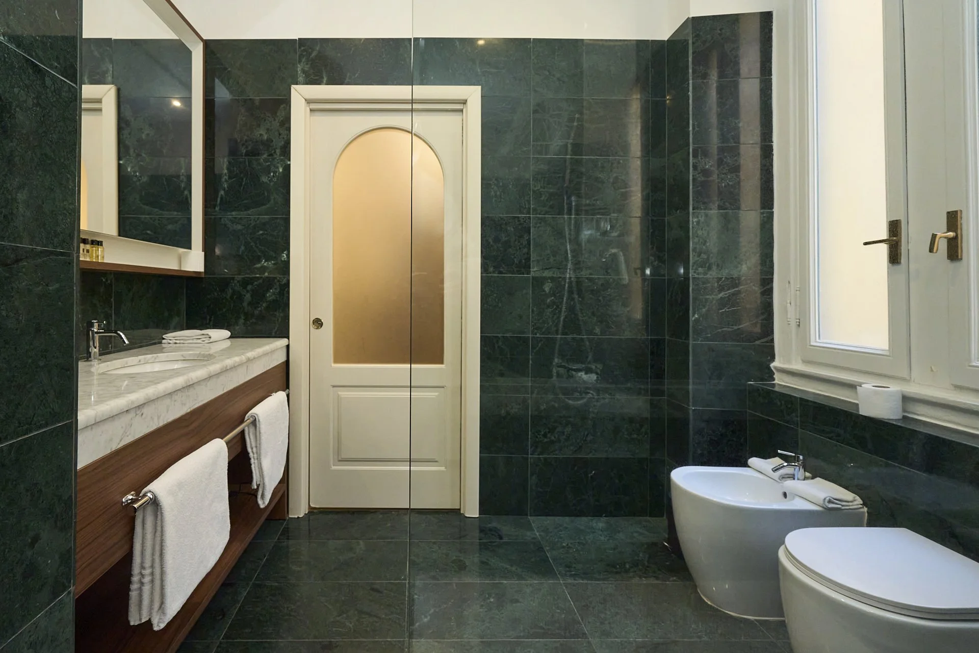 Bagno con pareti e pavimenti in marmo verde scuro, lavabo con mensola in marmo e due asciugamani bianchi, bidet e toilette bianco, finestra con persiana chiusa, e porta bianca col vetro opaco.