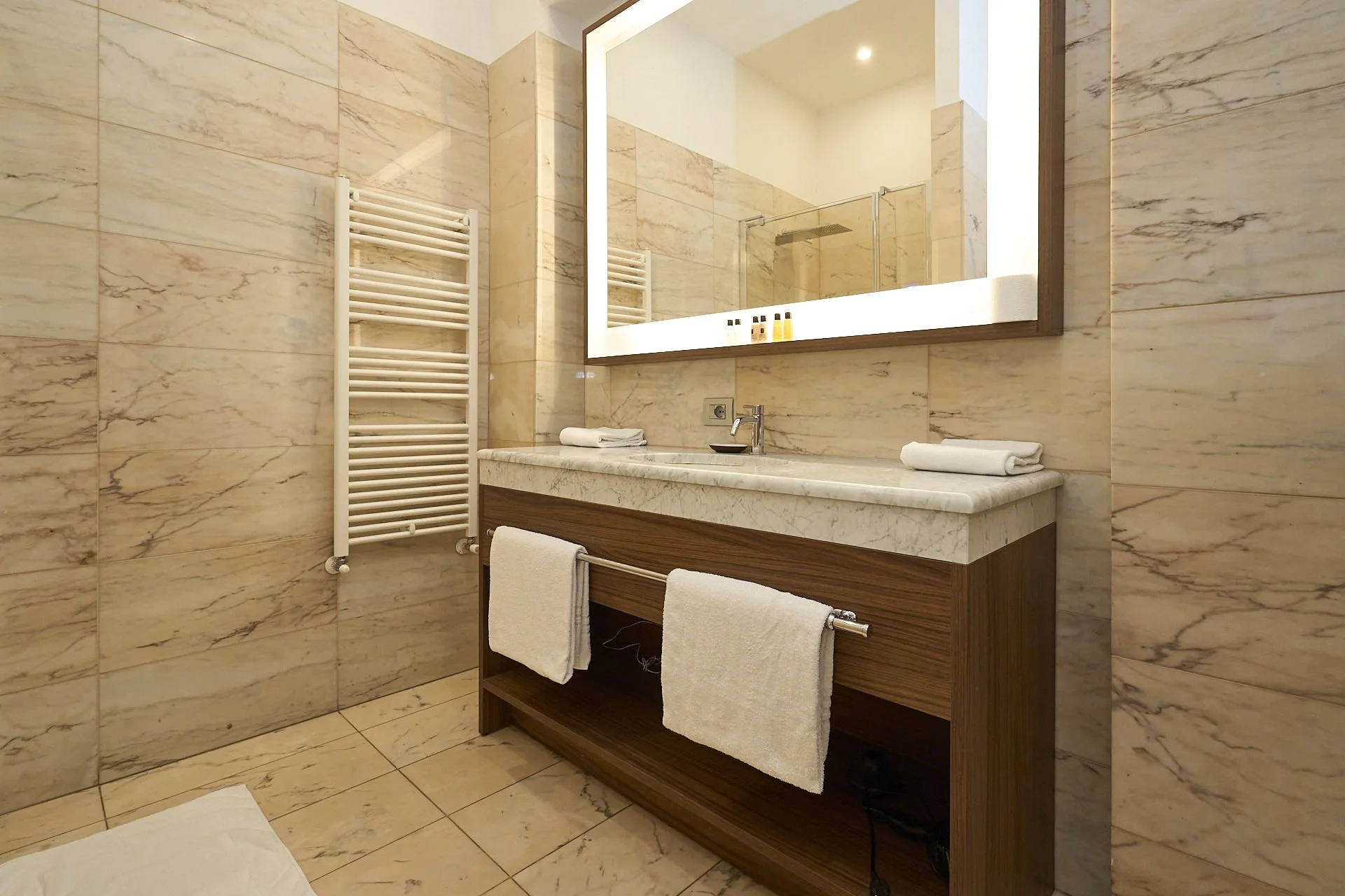 Bagno con pareti e pavimenti in marmo, grande specchio con luce frontale, lavabo su mobile in legno, asciugamani bianchi, radiator scaldasalviette, doccia visibile nello specchio.