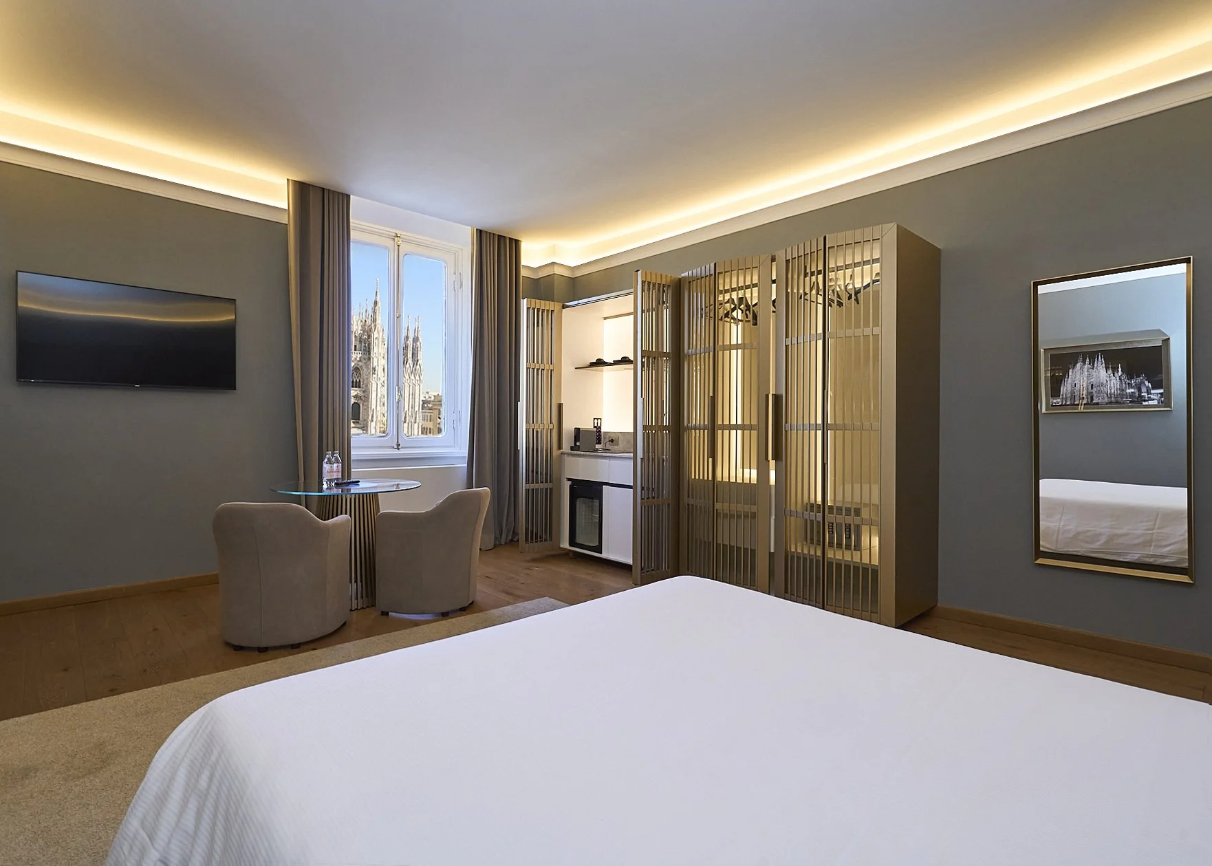 Interno di una camera d'albergo con letto bianco, pareti grigie, finestra con vista sulla cattedrale, TV a parete, specchio e area soggiorno con due sedie e tavolo.