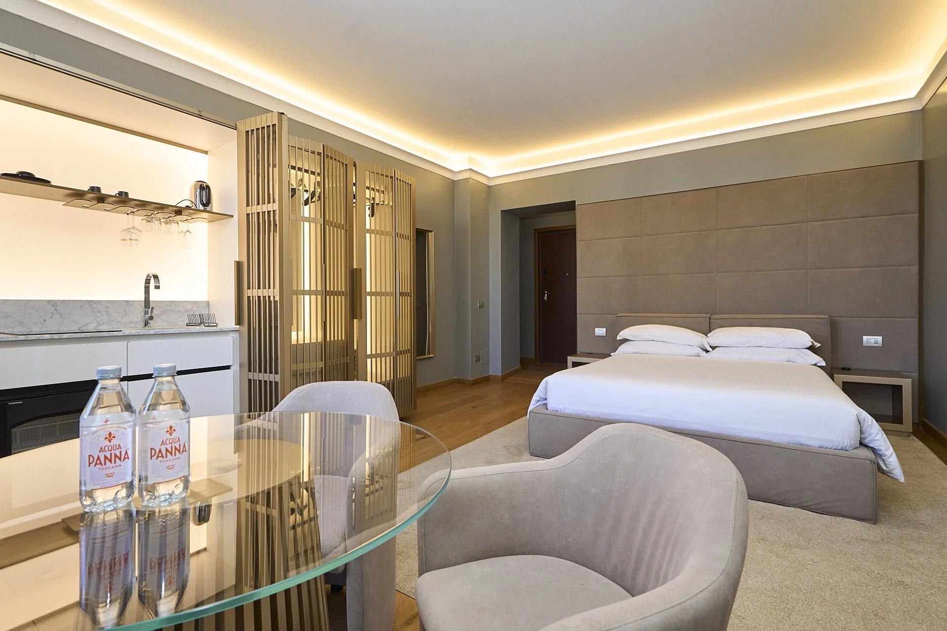 Camera da letto moderna con letto matrimoniale, pareti neutre, illuminazione soffusa e piccolo angolo con due bottiglie di acqua su un tavolo di vetro.