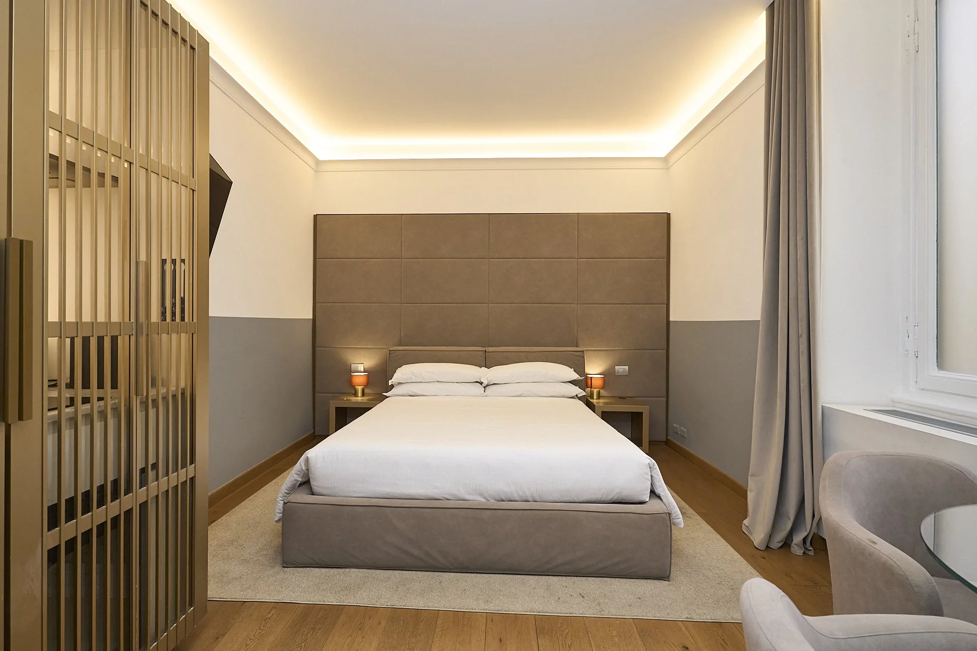 Camera matrimoniale elegante con letto, due comodini con lampade, grande testata imbottita, finestra con tende, sedie e tavolo.