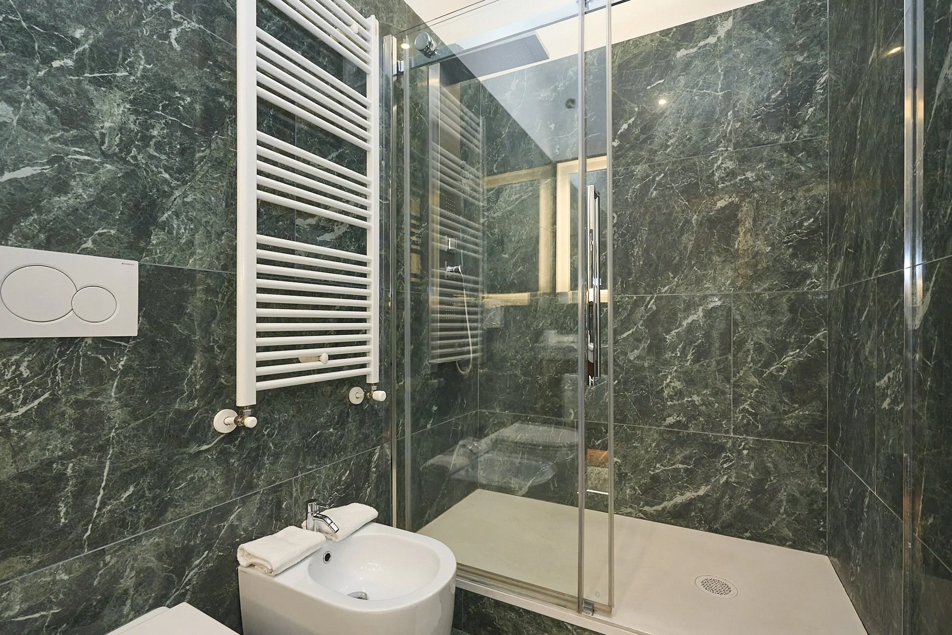 Bagno con pareti in marmo verde, doccia con porta a vetri e piatto doccia bianco, radiatore orizzontale bianco, bidet bianco con asciugamano arrotolato sopra, soffitto con finestra o lucernario sul soffitto