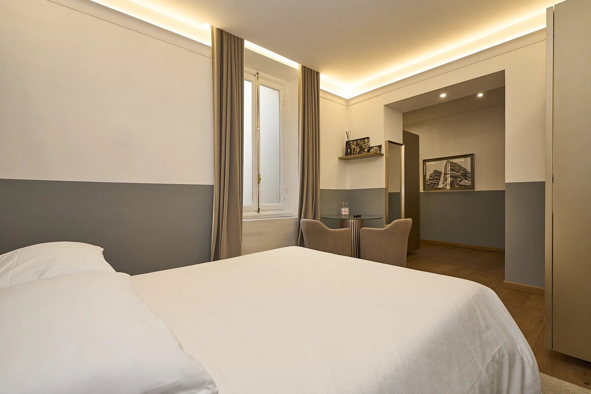 Camera da letto minimalista con letto bianco, finestra con tende beige, pareti bianche con area grigia, sedia e tavolino con acqua e snack, decori in stile moderno.