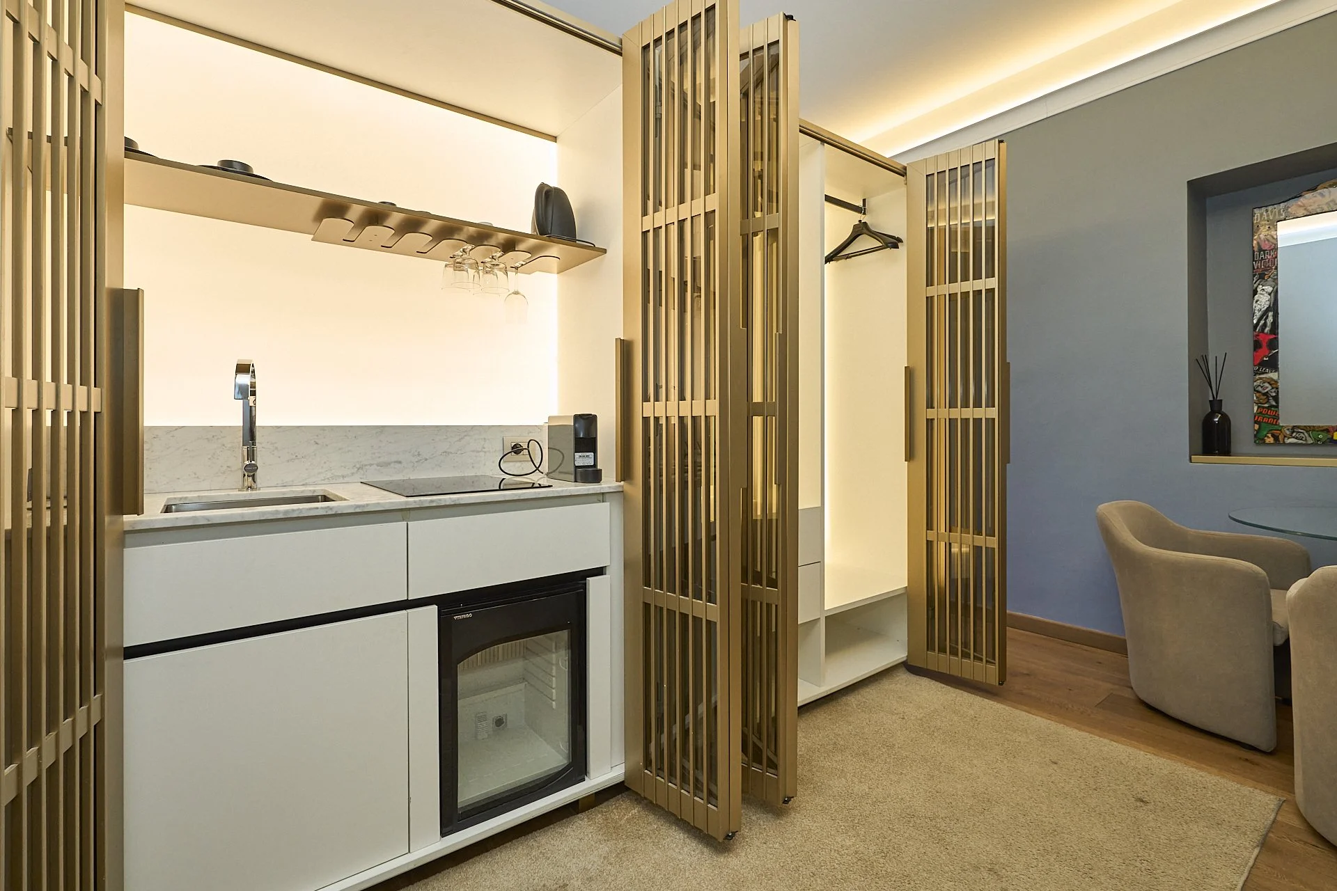 Mini cucina con ripiani in marmo, elettrodomestici, tè e bicchieri, divisori di legno, area soggiorno con sedie e tavolo.