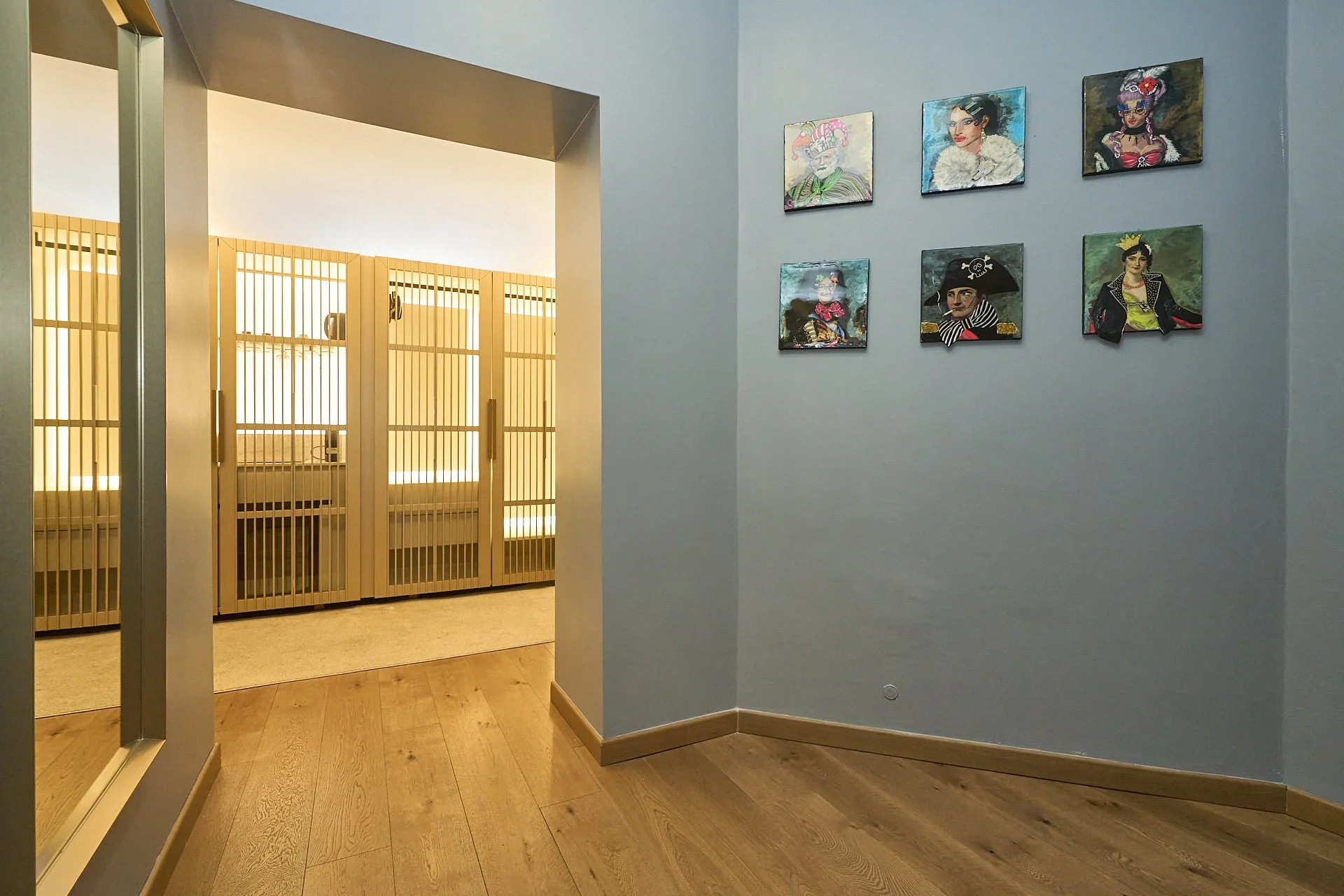 Muro con sei quadri di stile diverso, parete azzurra, pavimento in legno, ingresso a un'area con gabbie in legno e vetro.