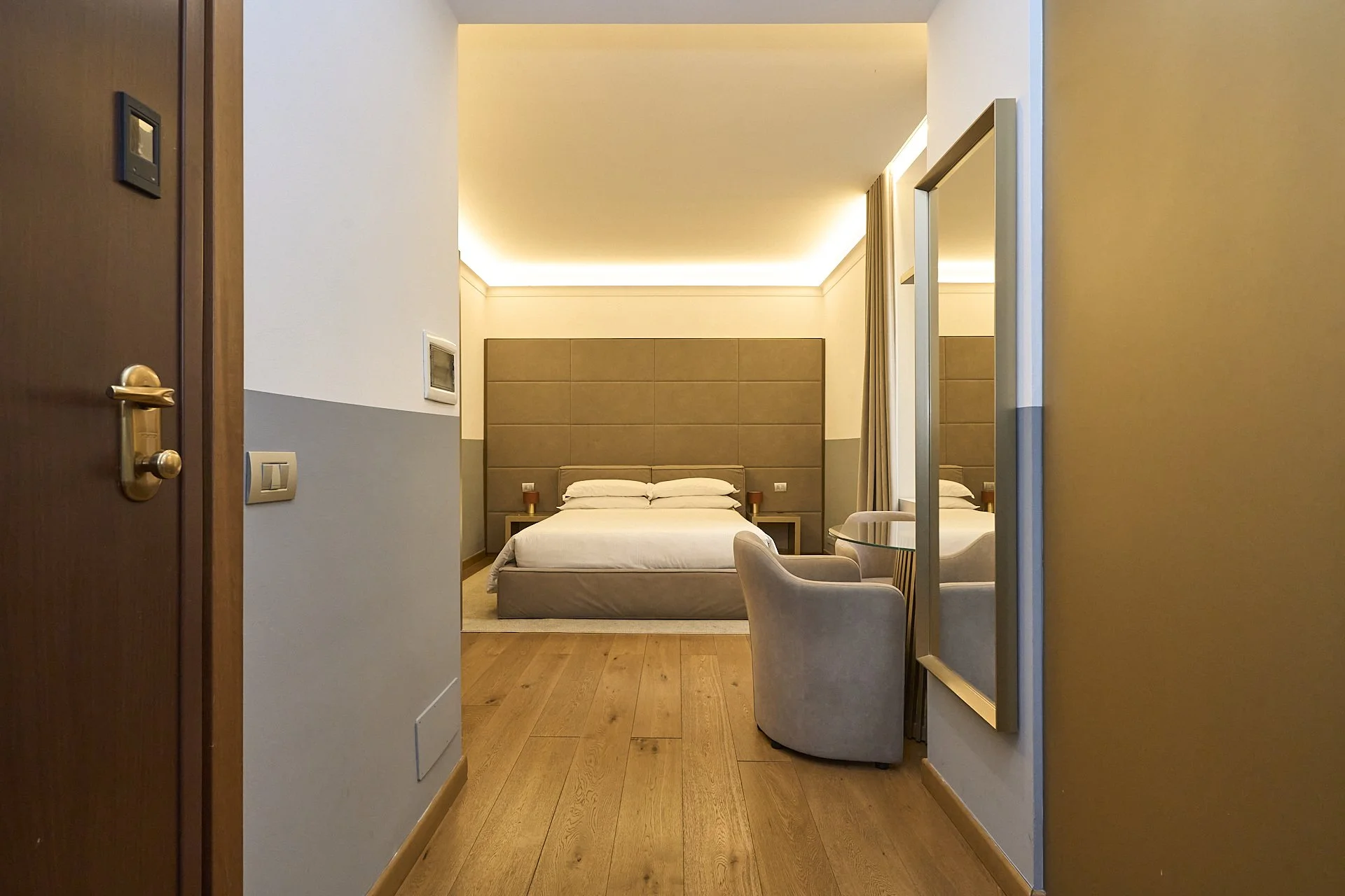 Vista di una camera da letto moderna e ben arredata, con letto matrimoniale, poltrone, specchio e luci soffuse.