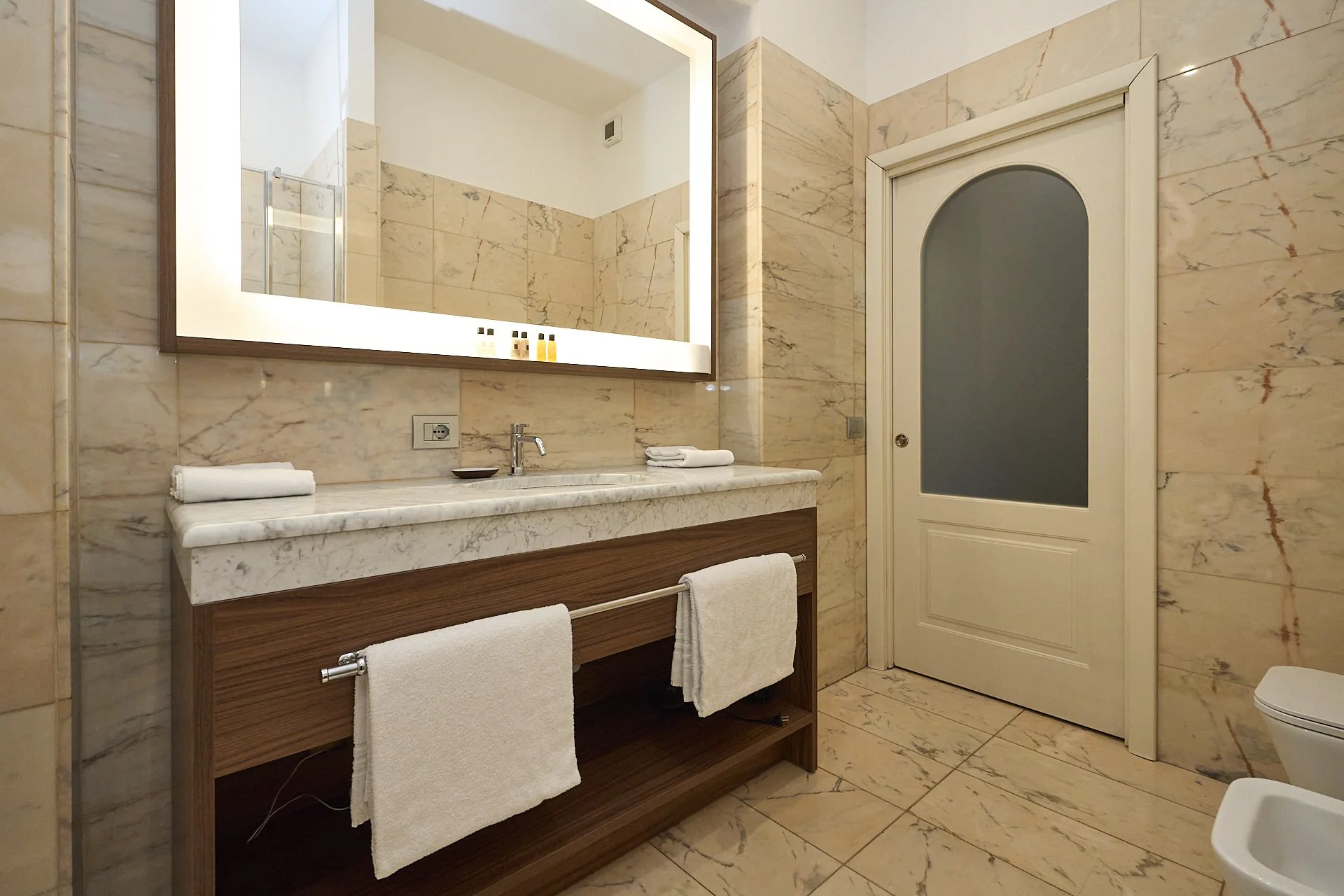 Bagno con pareti e pavimenti in marmo beige, grande specchio con illuminazione, lavabo con superficie in marmo, asciugamani bianchi, porta bianca con ante in vetro satinato, bidet visibile in parte.