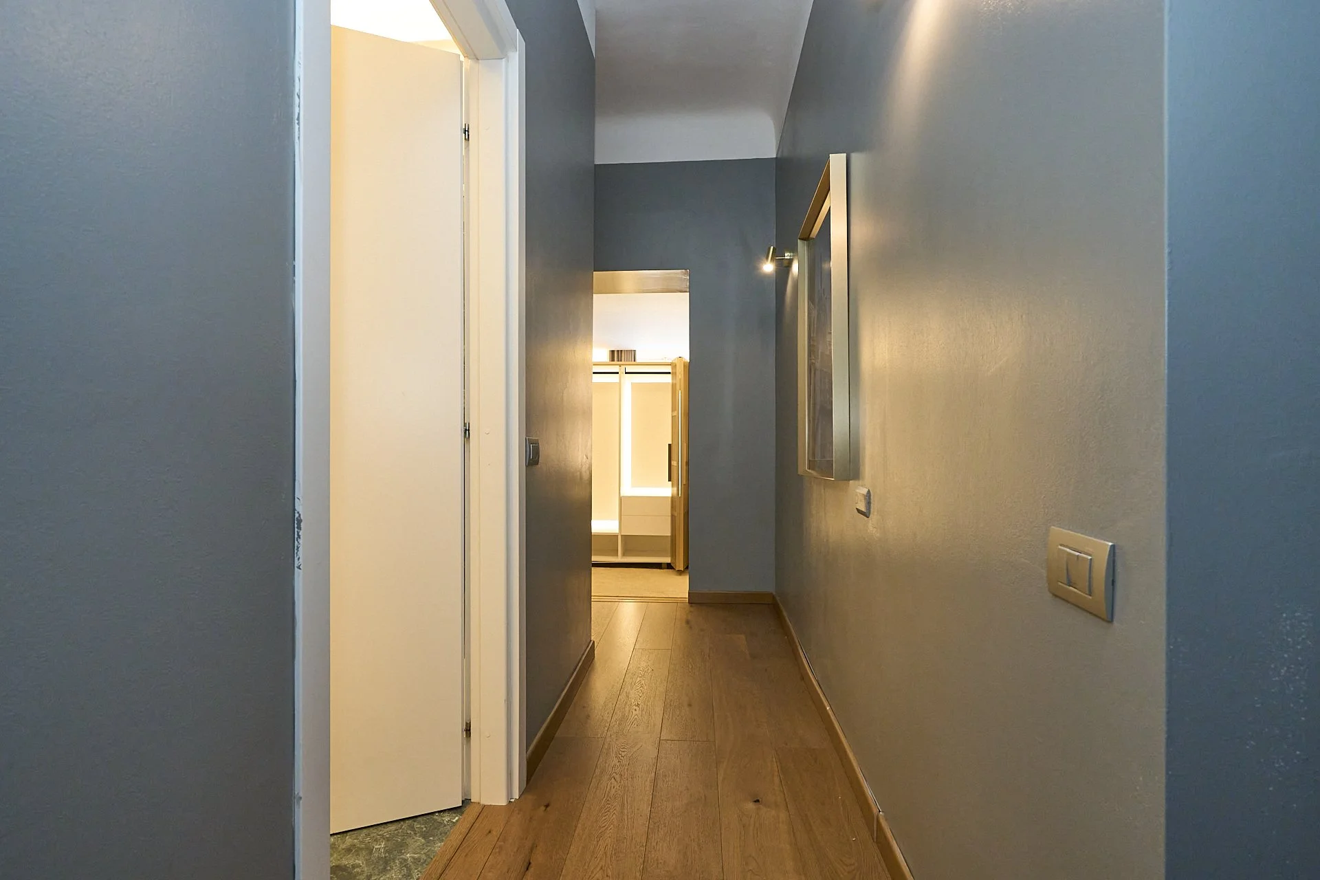 Ingresso di un corridoio di una casa con pareti blu, un pavimento di legno e una porta bianca aperta che conduce a una stanza con interni chiari.