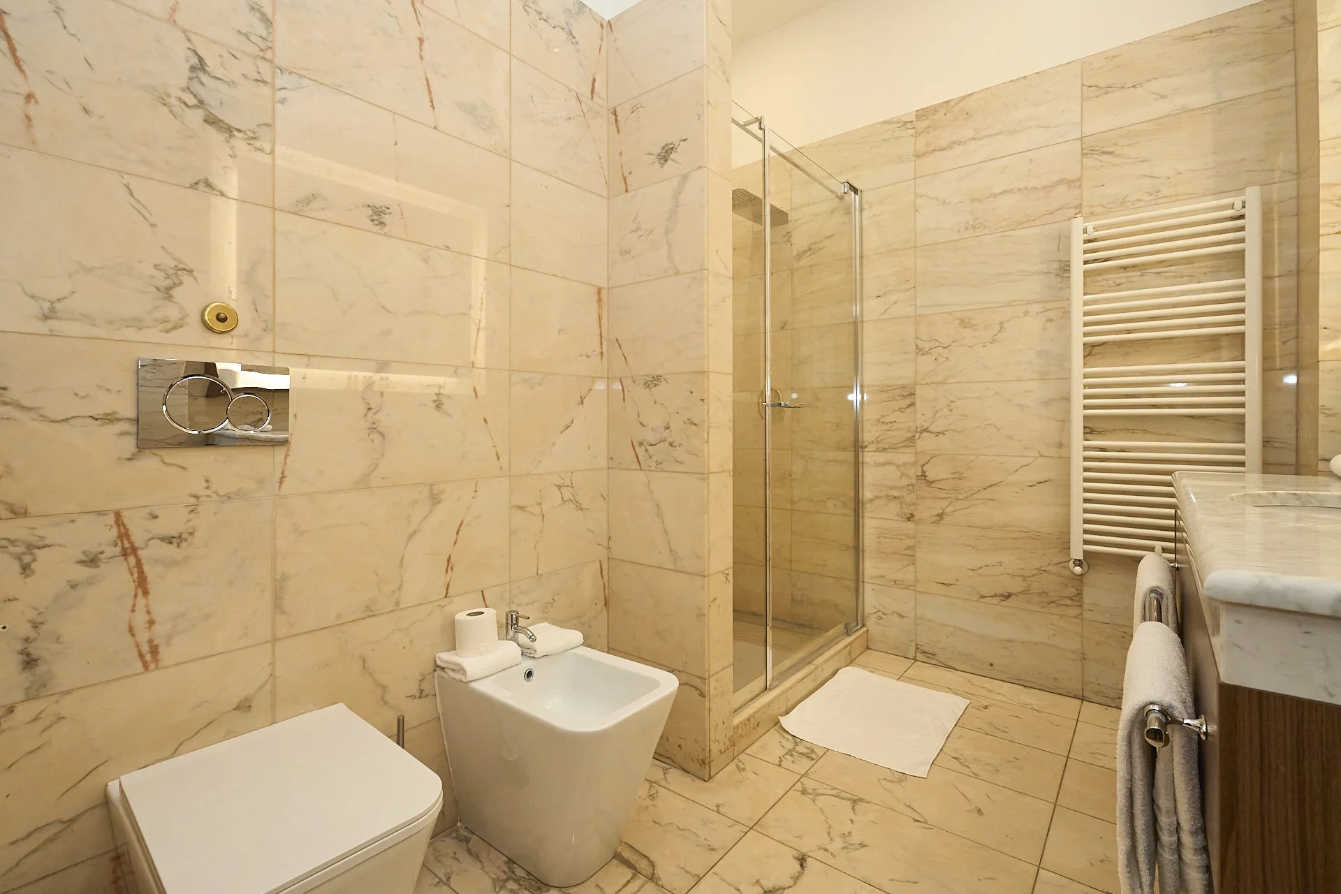 Bagno con pareti e pavimento in marmo crema, doccia con porta in vetro, lavabo in marmo con asciugamani, radiatore bianco, bidet, wc, specchio con acciaieria