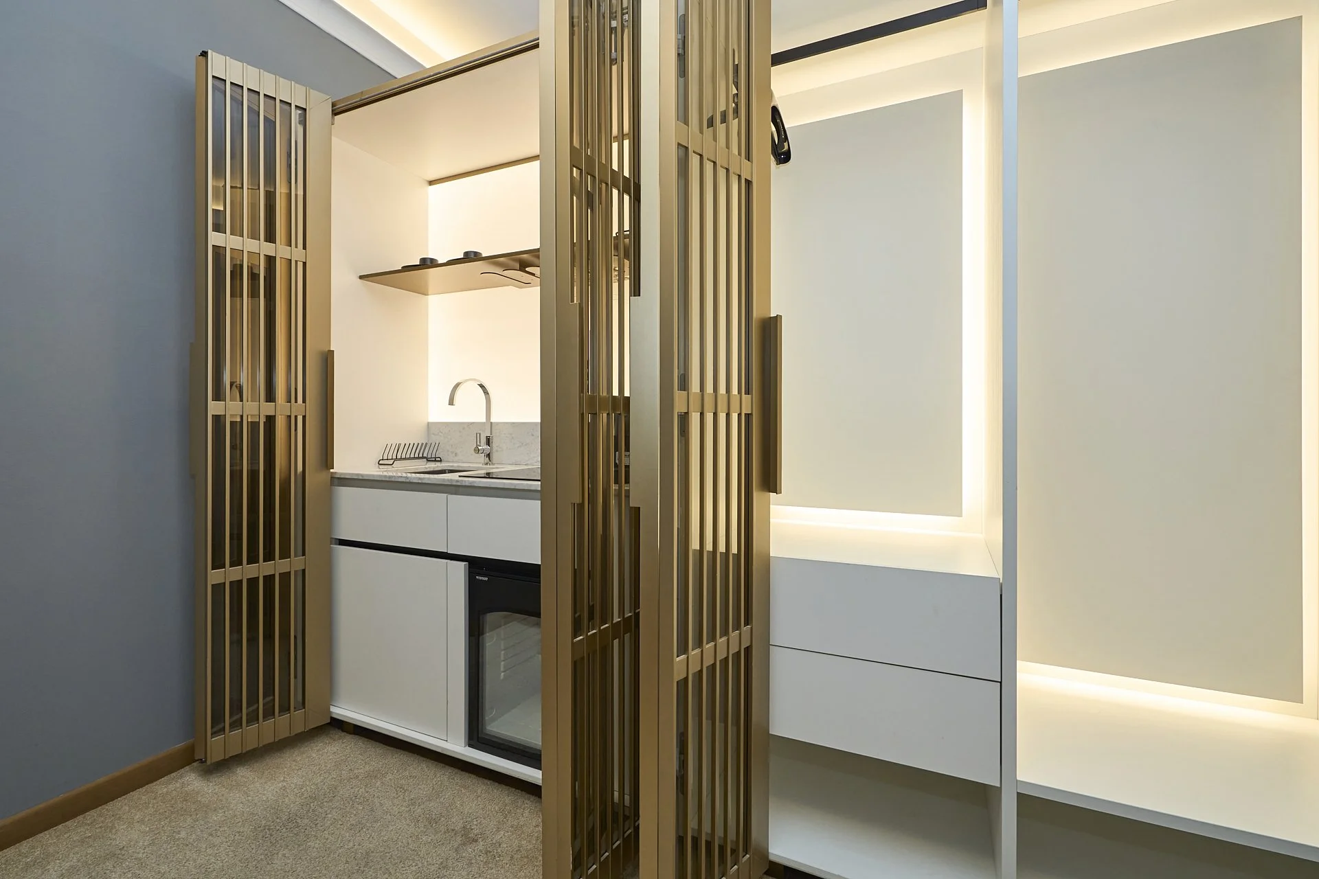 Angolo cucina con frigorifero sotto il lavabo in un ambiente moderno, separato da pannelli di legno open-frame.