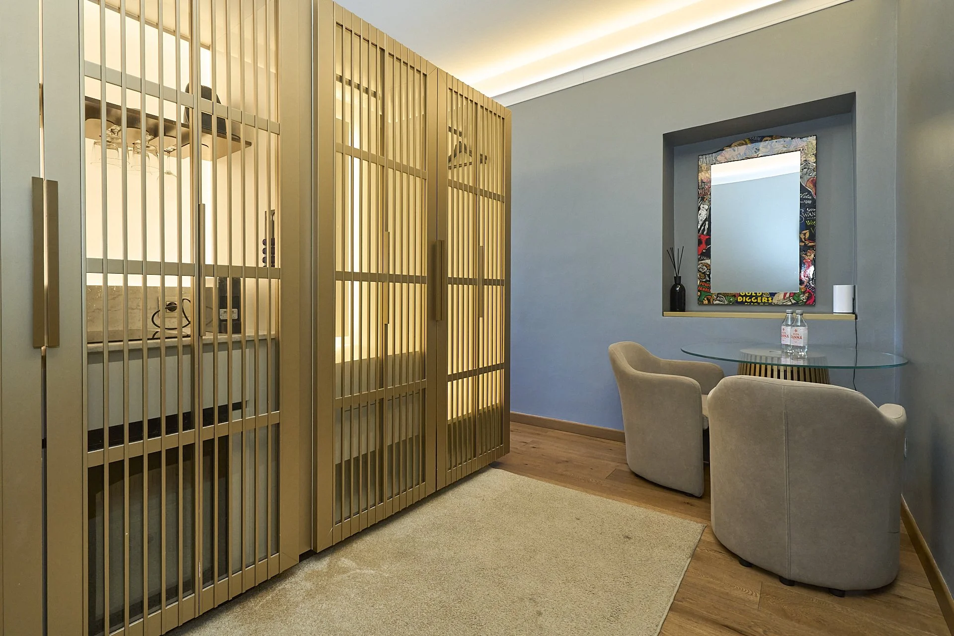 Interno di una stanza con parete blu, due sedie beige e un tavolino di vetro, decorato con un quadro colorato, una bottiglia d'acqua e un diffusore di aromi, con un’anta scorrevole in metallo dorato e una vaschetta con bicchieri e bottiglia di vino.