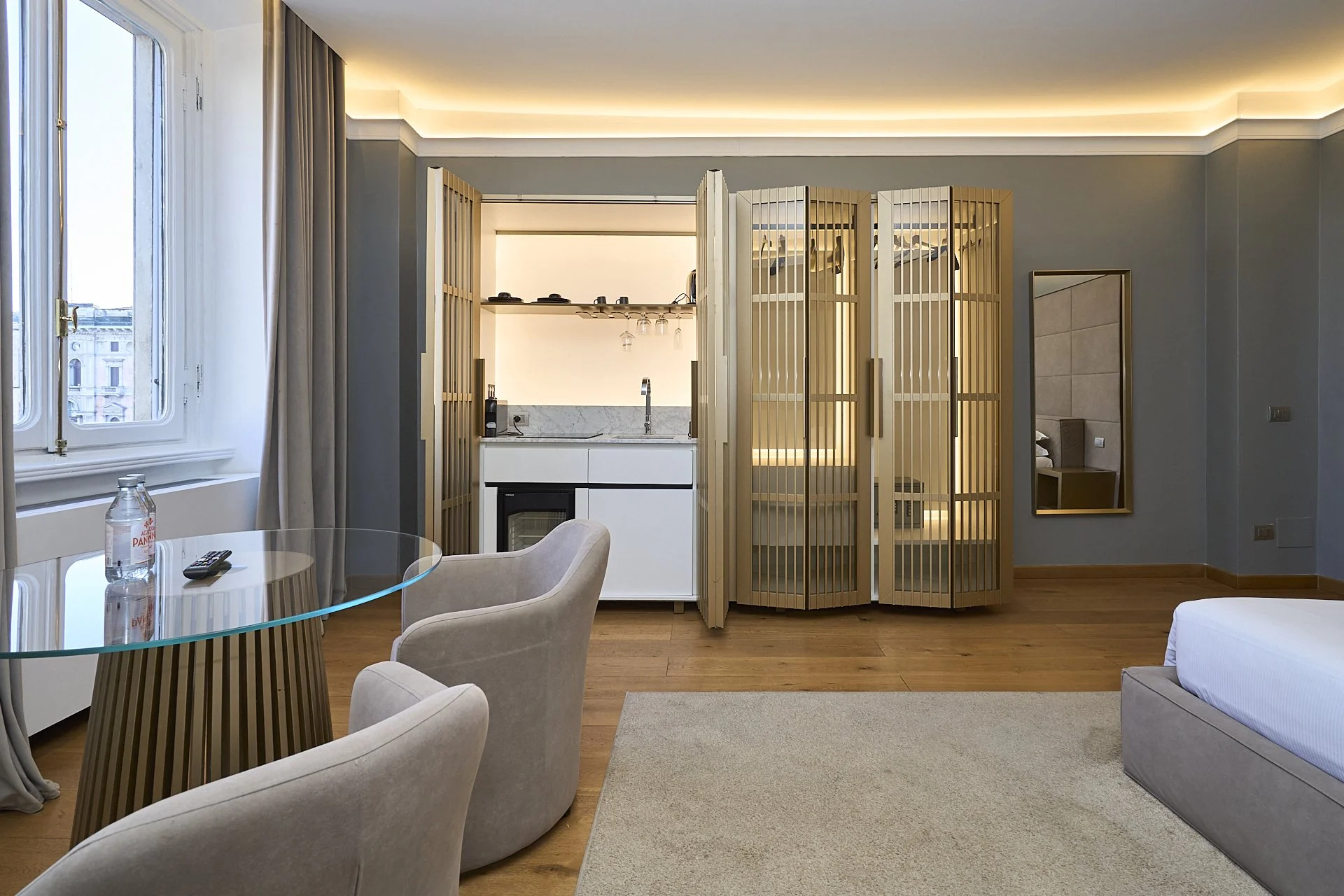 Interno di una camera d'hotel con zona soggiorno, tavolo di vetro, sedie, finestra, cucina compatta con divisorio in legno e porta, specchio e letto parzialmente visibile.