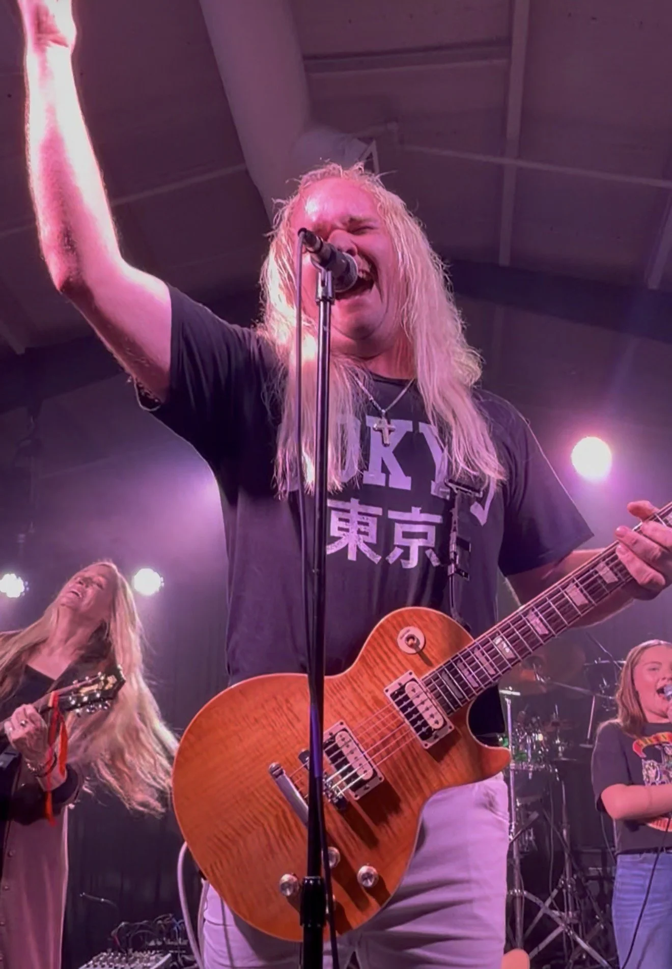 A rock musician with long blond hair and a black T-shirt singing passionately into a microphone while playing an orange electric guitar on a stage with purple lighting.