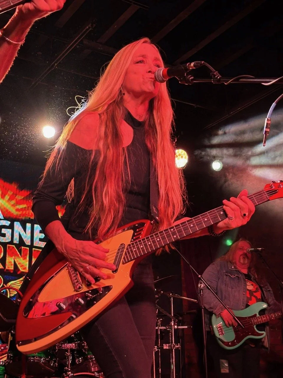 A female musician with long blonde hair singing into a microphone and playing an electric guitar on stage, with a male guitarist in the background, during a live music performance.