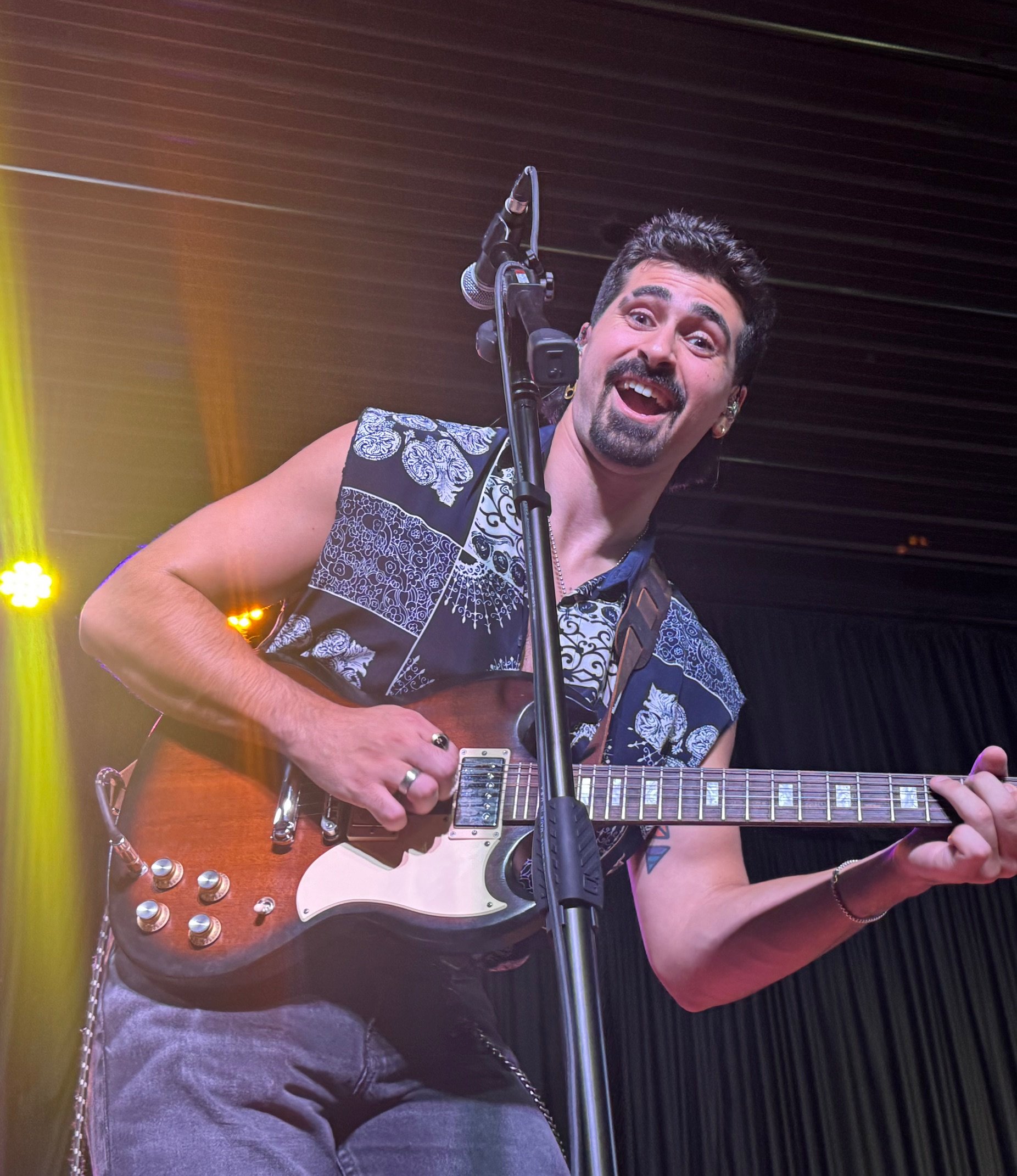A man with dark hair, a beard, and a mustache playing an electric guitar on stage, smiling and looking at the camera, with colorful stage lights in the background.