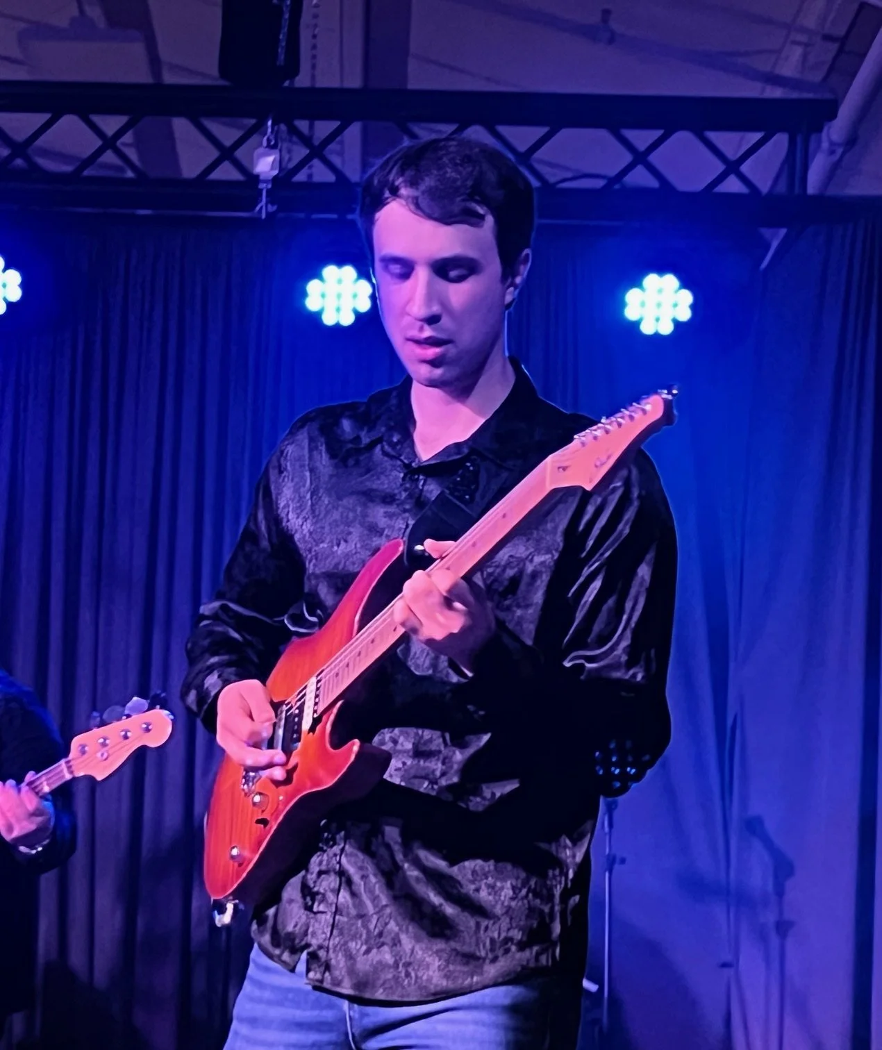 A man playing an electric guitar on stage under blue lighting with a black curtain backdrop.