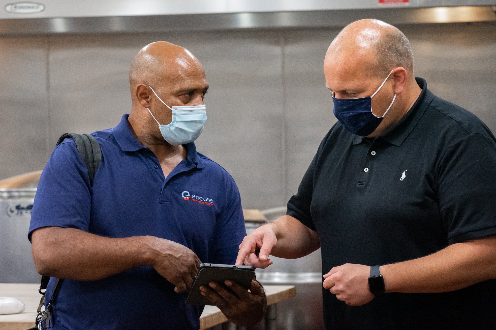 Two men wearing face masks discussing work on a tablet in a commercial kitchen.