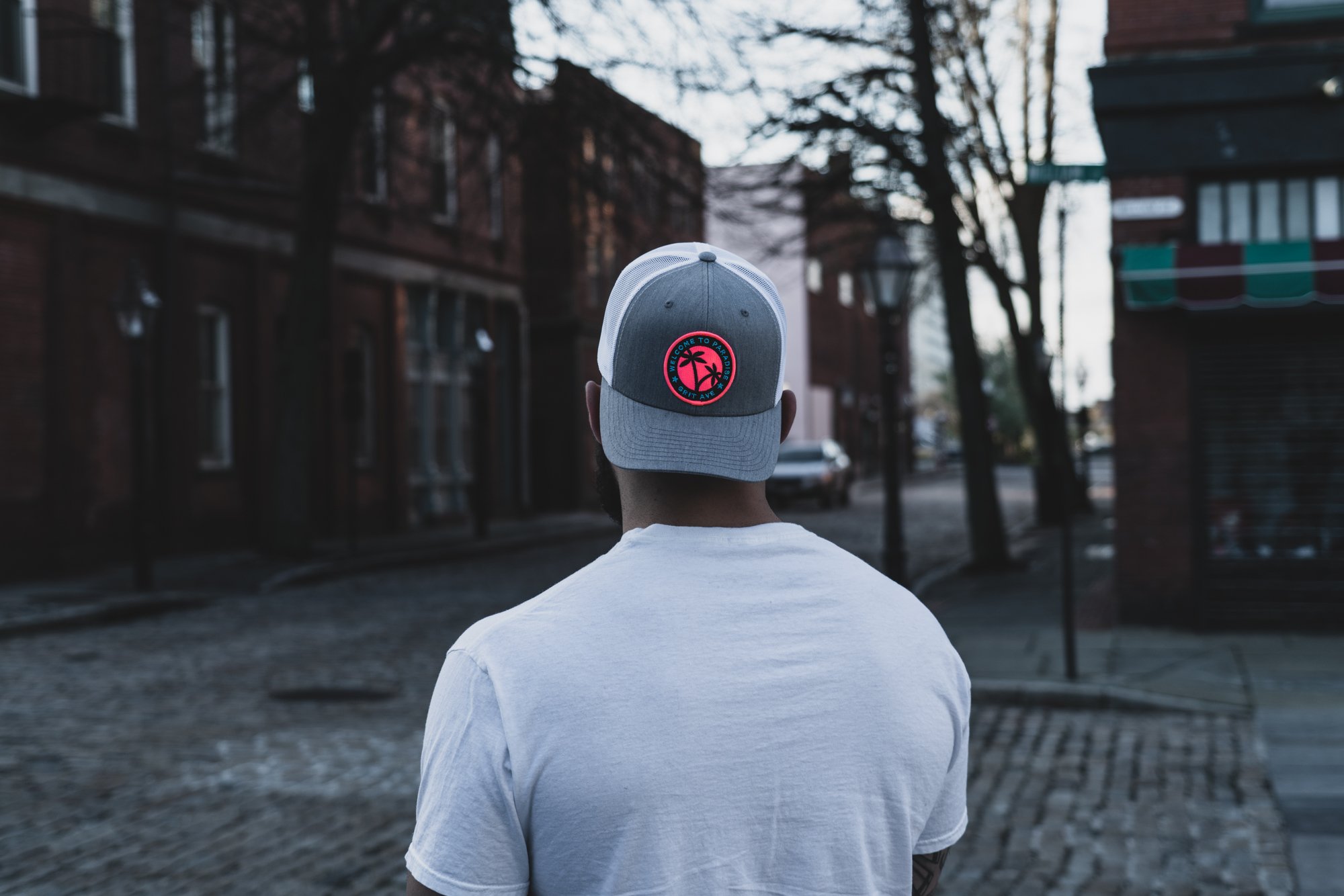 Man in white t-shirt and gray baseball cap standing on cobblestone street, urban setting.