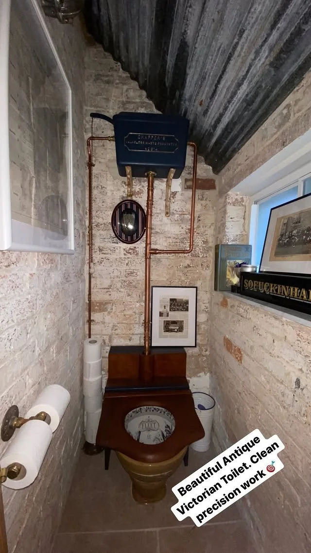 Antique Victorian toilet with a dark wooden seat and a large copper flush tank mounted on the wall, with exposed brick walls and vintage decorations.