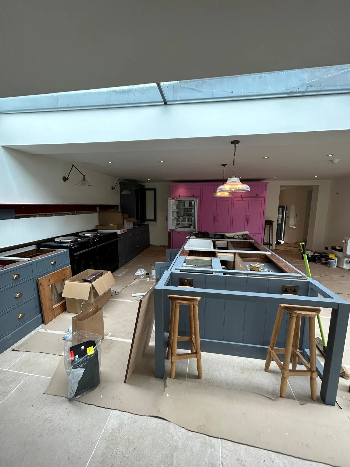 Kitchen under renovation with unfinished cabinetry, tools, and construction materials, including a pink cabinet, blue island frame, and stools.