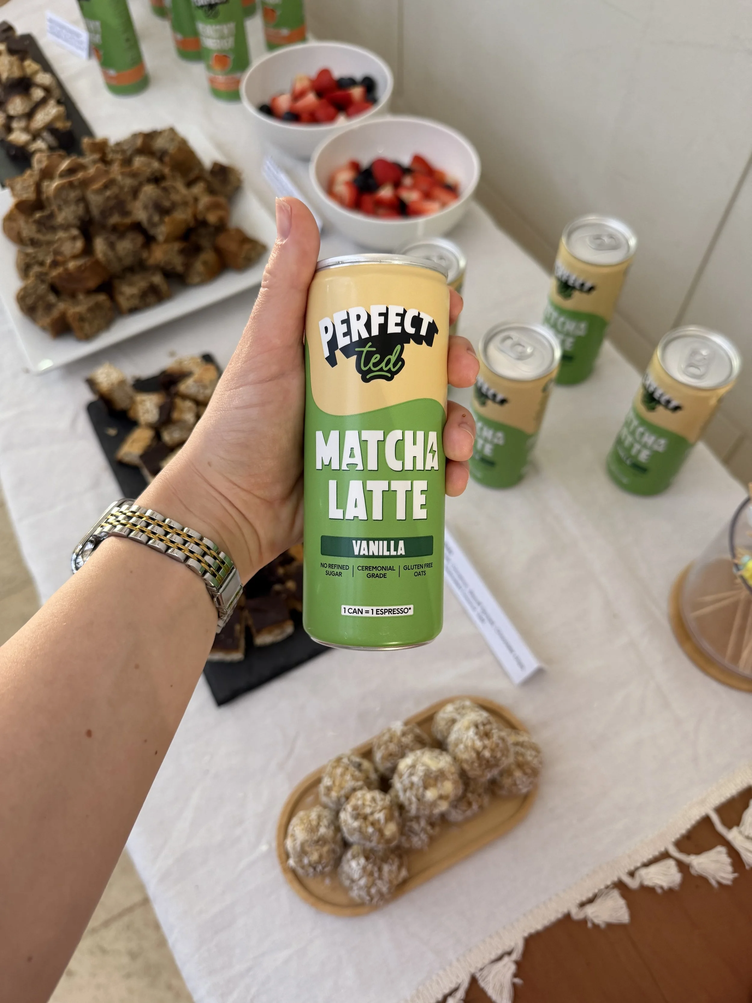 A hand holding a can of Perfect Ted Matcha Latte Vanilla with multiple other cans on a table, alongside bowls of fruit salad and a tray of assorted pastries or baked goods.