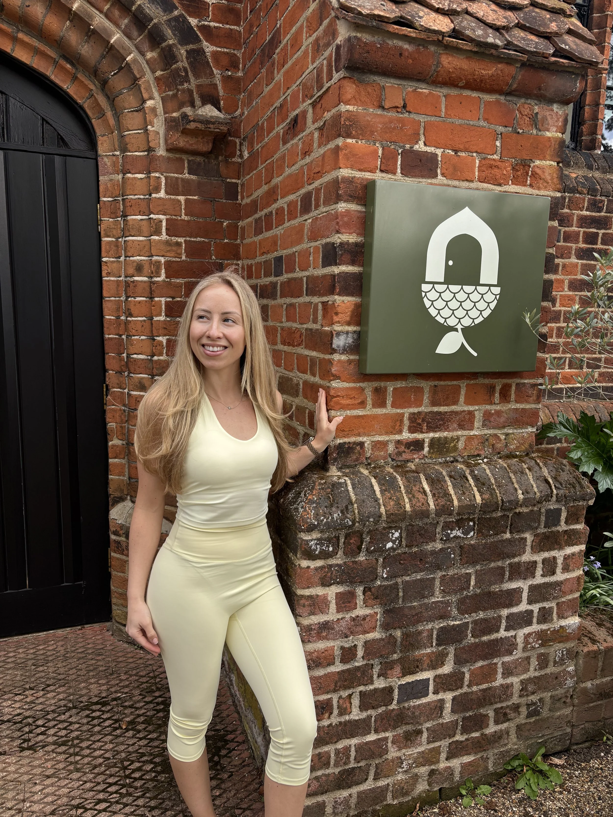 A woman with long blonde hair wearing a pale yellow dress stands outside near a brick wall with a decorative sign featuring an illustration of a door and fish scales. She is smiling and looking to her left.