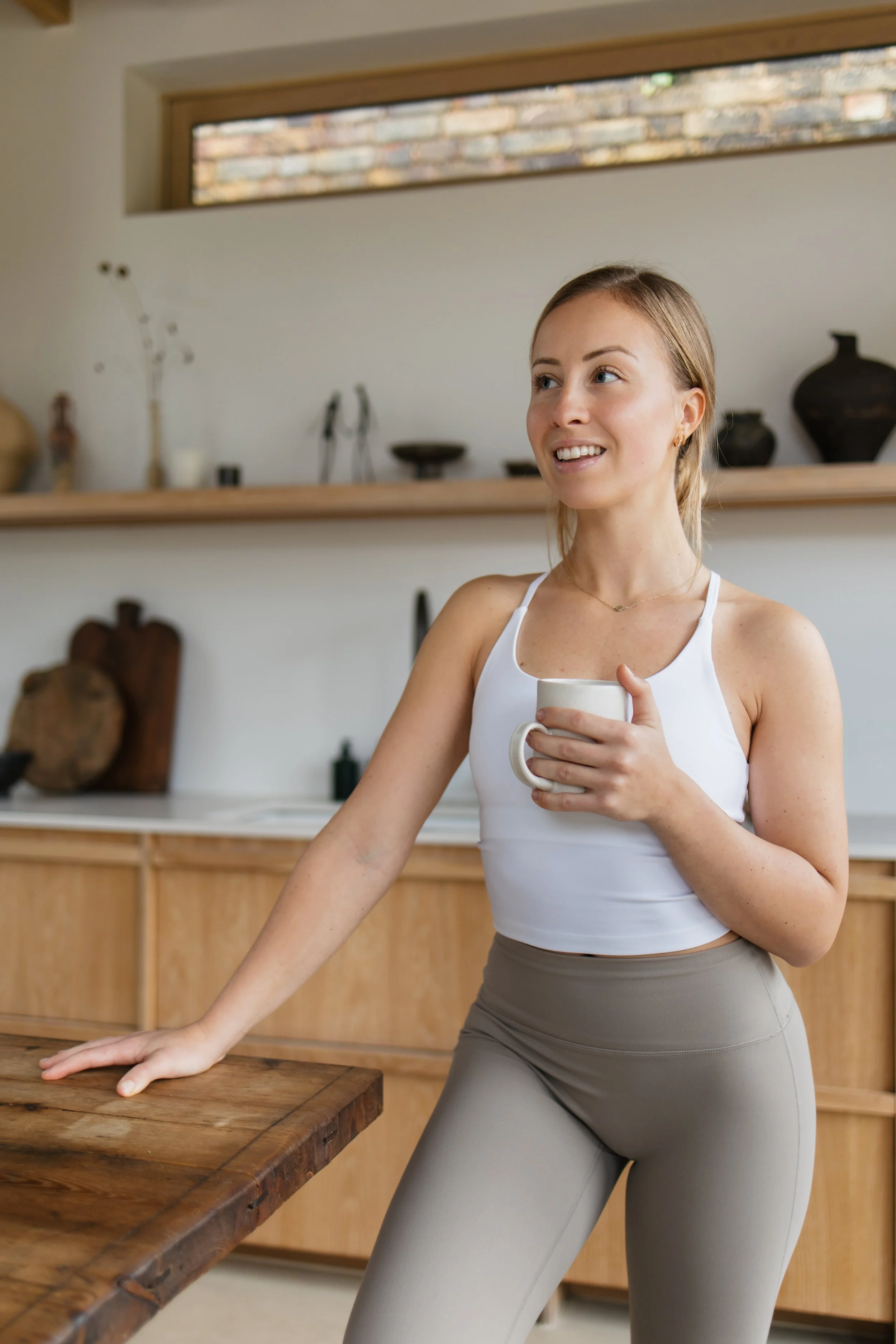 A woman with blonde hair, wearing a white tank top and gray leggings, leans on a wooden kitchen counter, holding a white mug and smiling. The background features a shelf with decorative items and a window showing a brick wall outside.