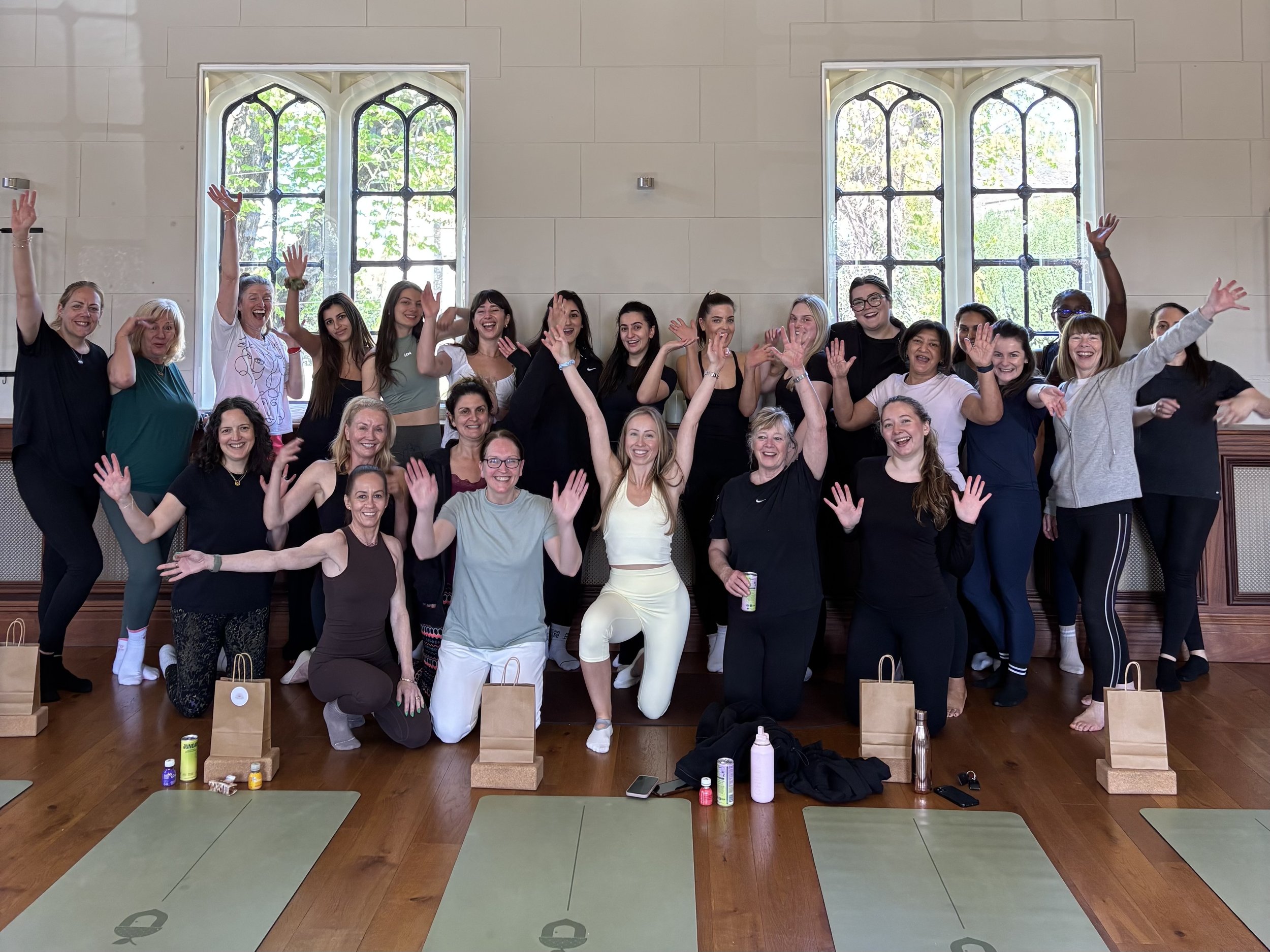 A large group of women gathered in a room, smiling and waving at the camera. The room has tall, ornate windows with greenery outside. The women are standing and kneeling in front of a wall, with yoga mats and small paper bags on the floor in front of them. Some have water bottles and personal items nearby. The atmosphere appears joyful and social.