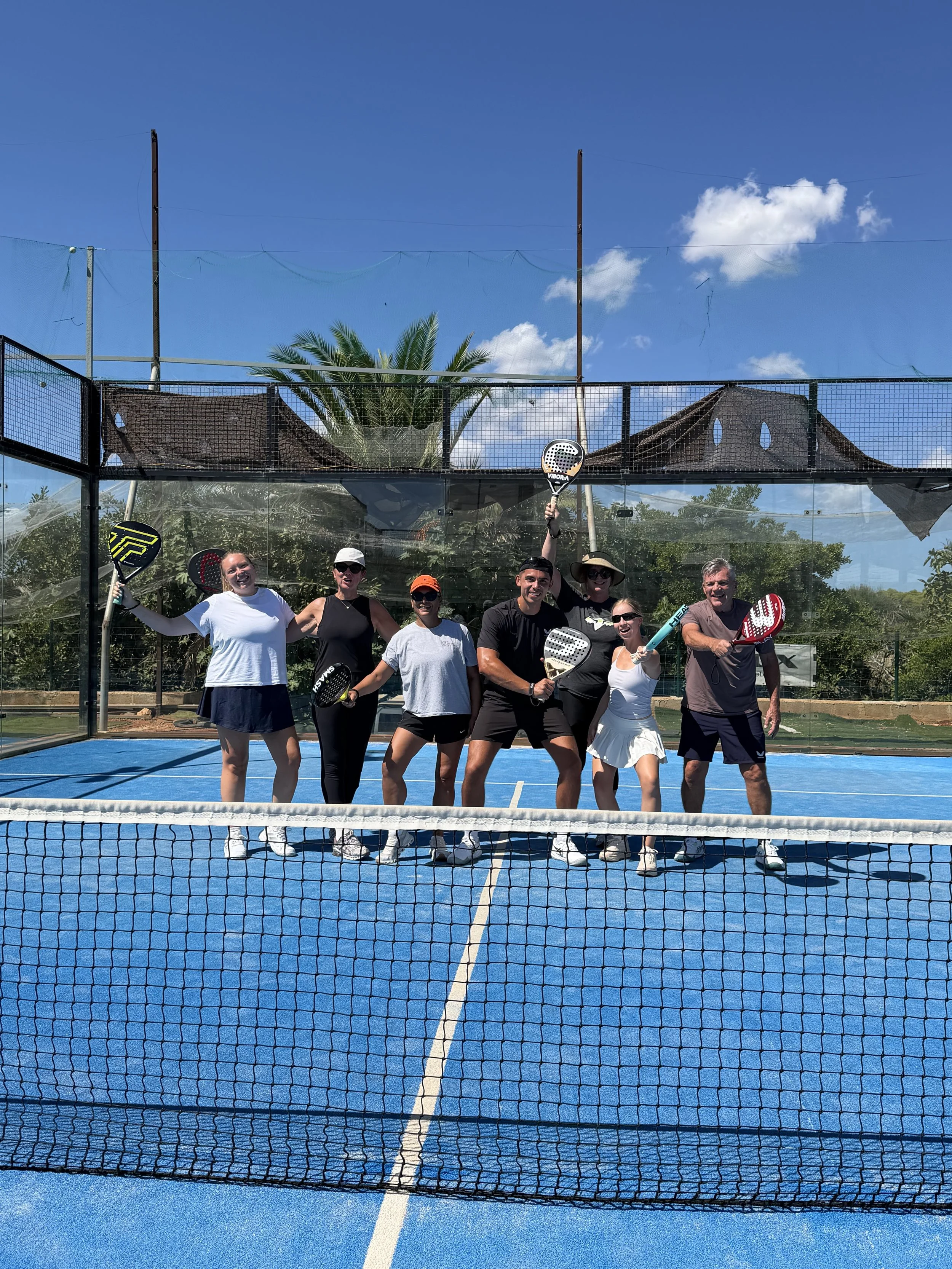 Group of six people smiling and holding padel rackets on a padel court with a net in the foreground, blue sky, palm trees, and nets in the background.
