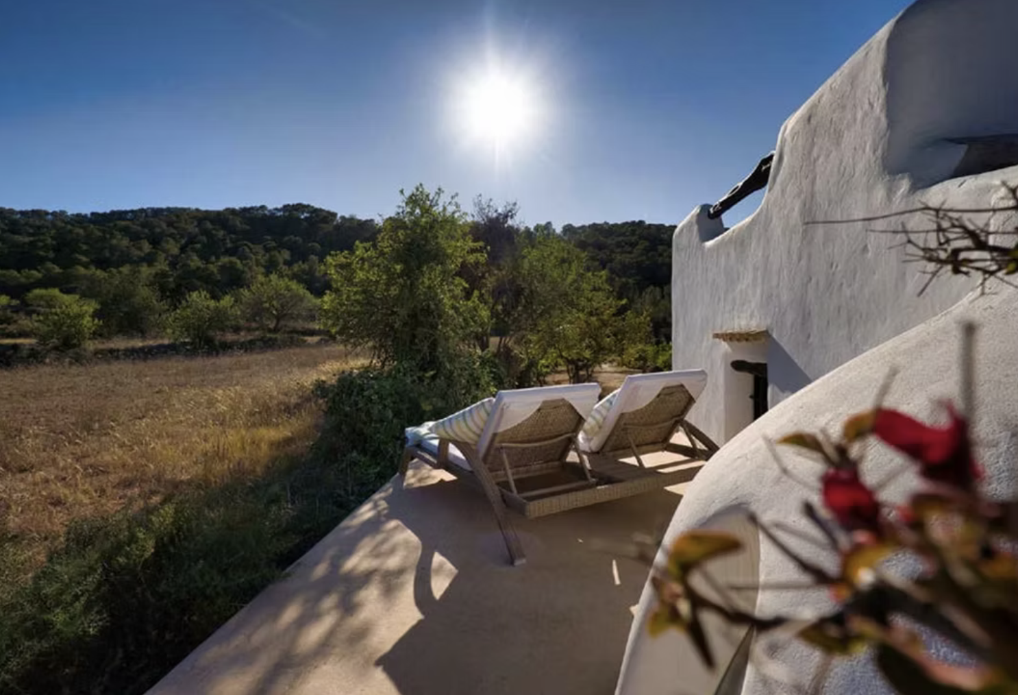 Sunlit terrace with two lounge chairs next to a white stucco wall, overlooking a landscape of trees and hills under a clear blue sky.