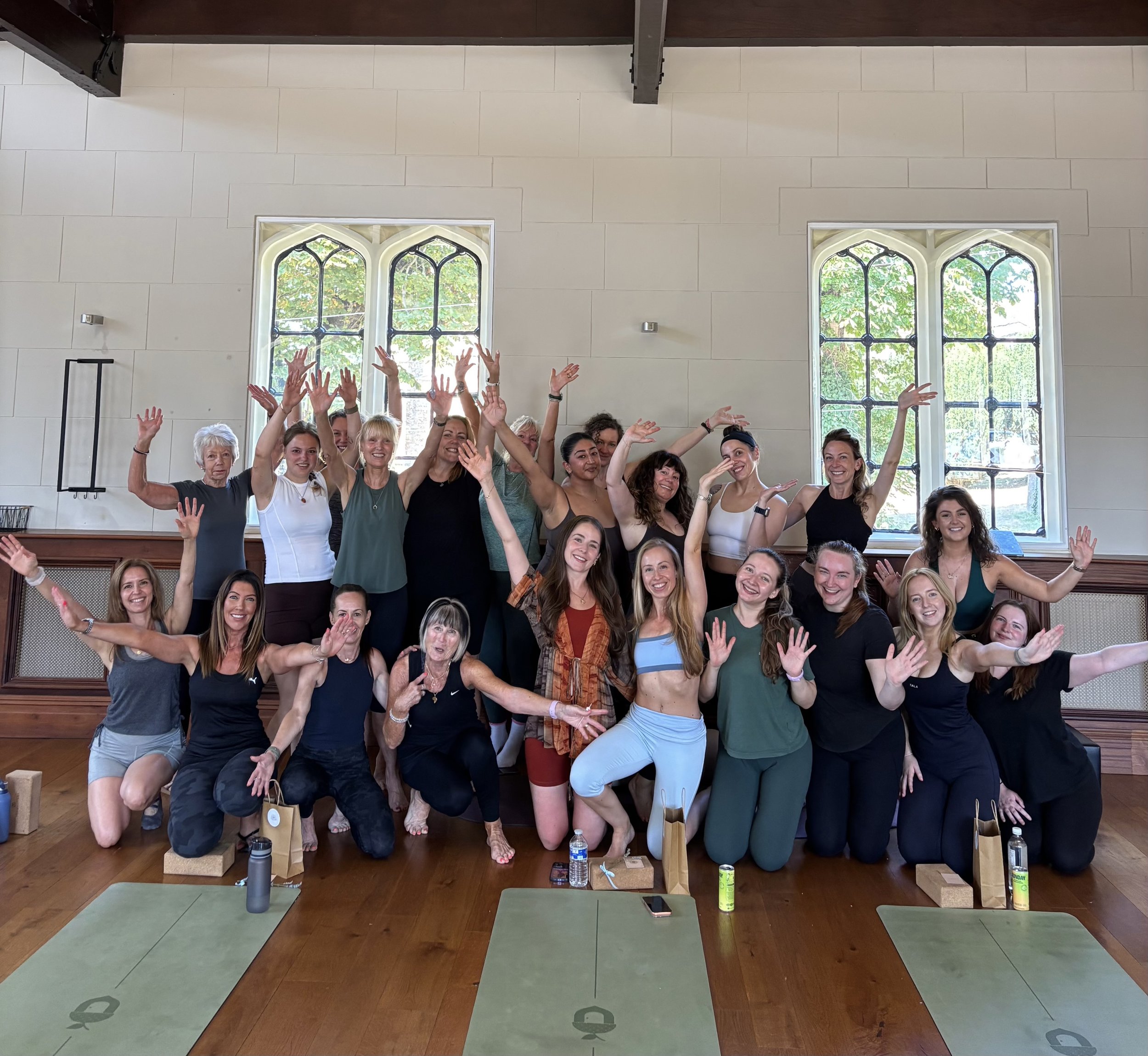 Group of diverse women participating in a yoga or meditation class indoors, some with raised hands, in front of large stained-glass windows. Yoga mats and water bottles are visible on the floor.