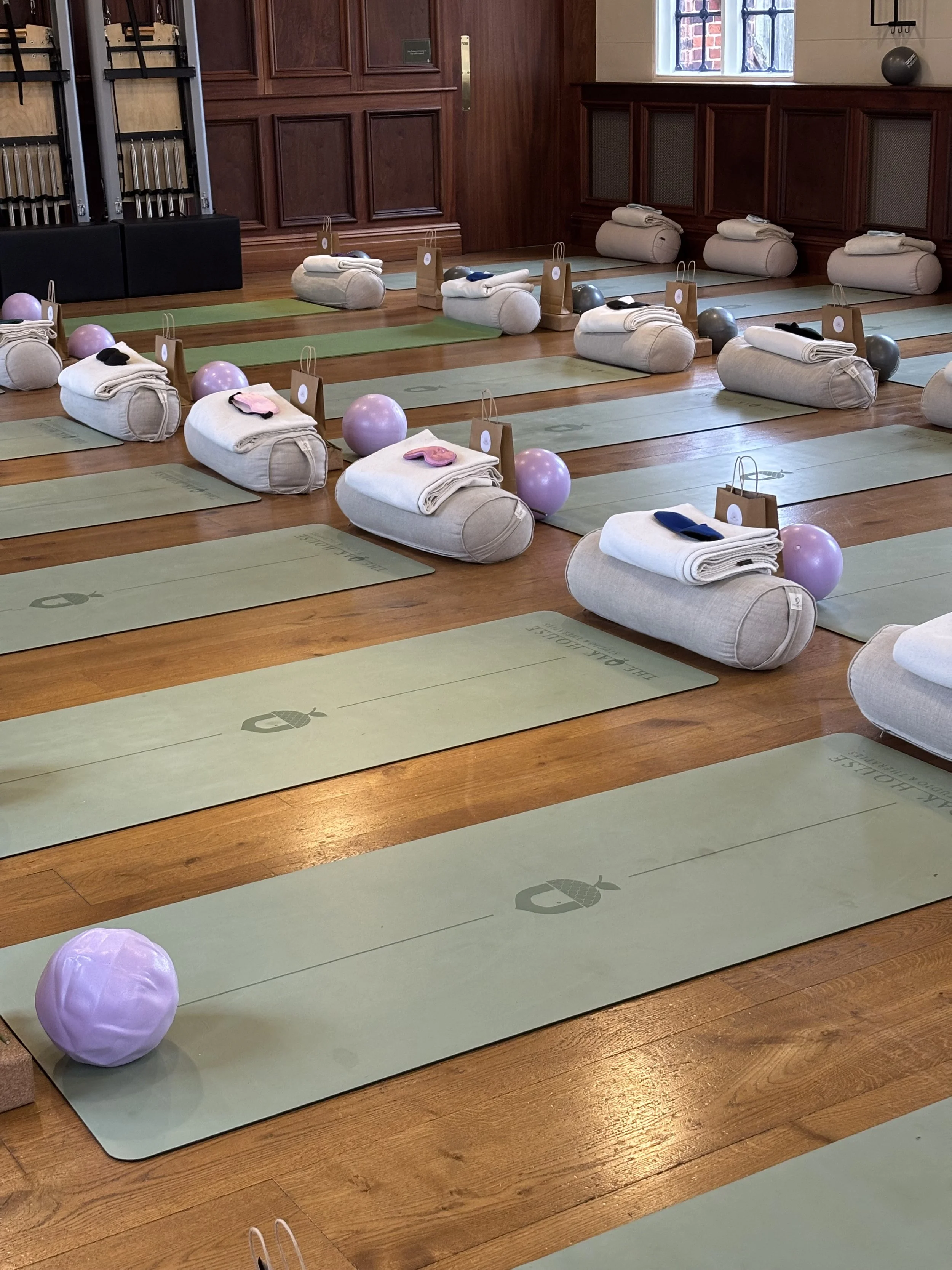 A yoga studio room prepared for a class, with yoga mats, rolled towels, small bags, pink exercise balls, and black eye masks laid out on the floor, with large windows and wooden paneling walls.