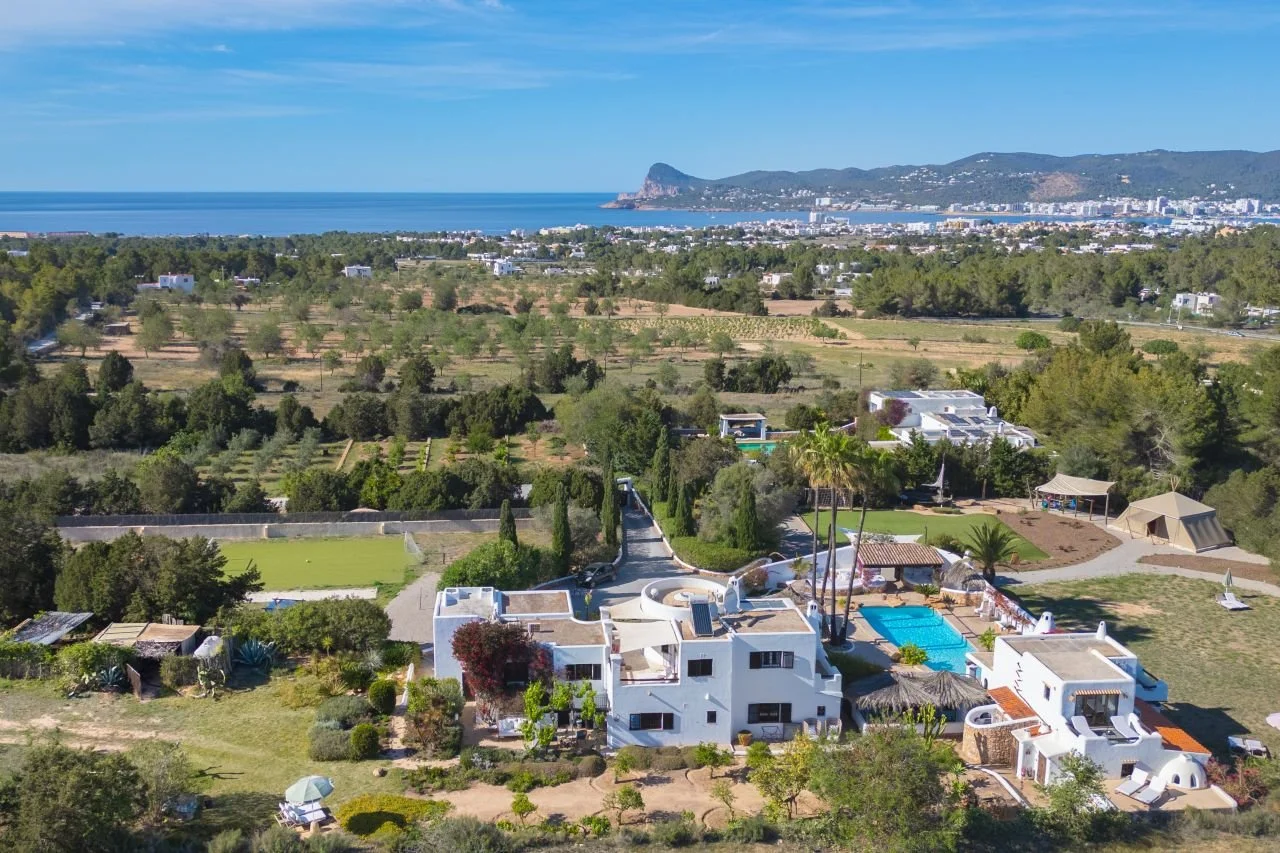 Aerial view of a luxurious white villa with swimming pool, surrounded by lush greenery and trees, with a coastline and cityscape in the background.