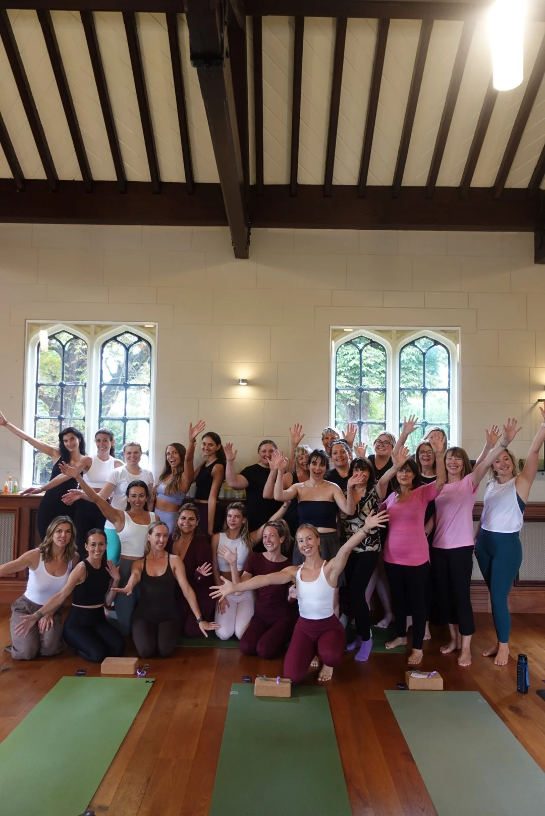 Group of women participating in a yoga class in a bright studio with tall windows and wooden floors.