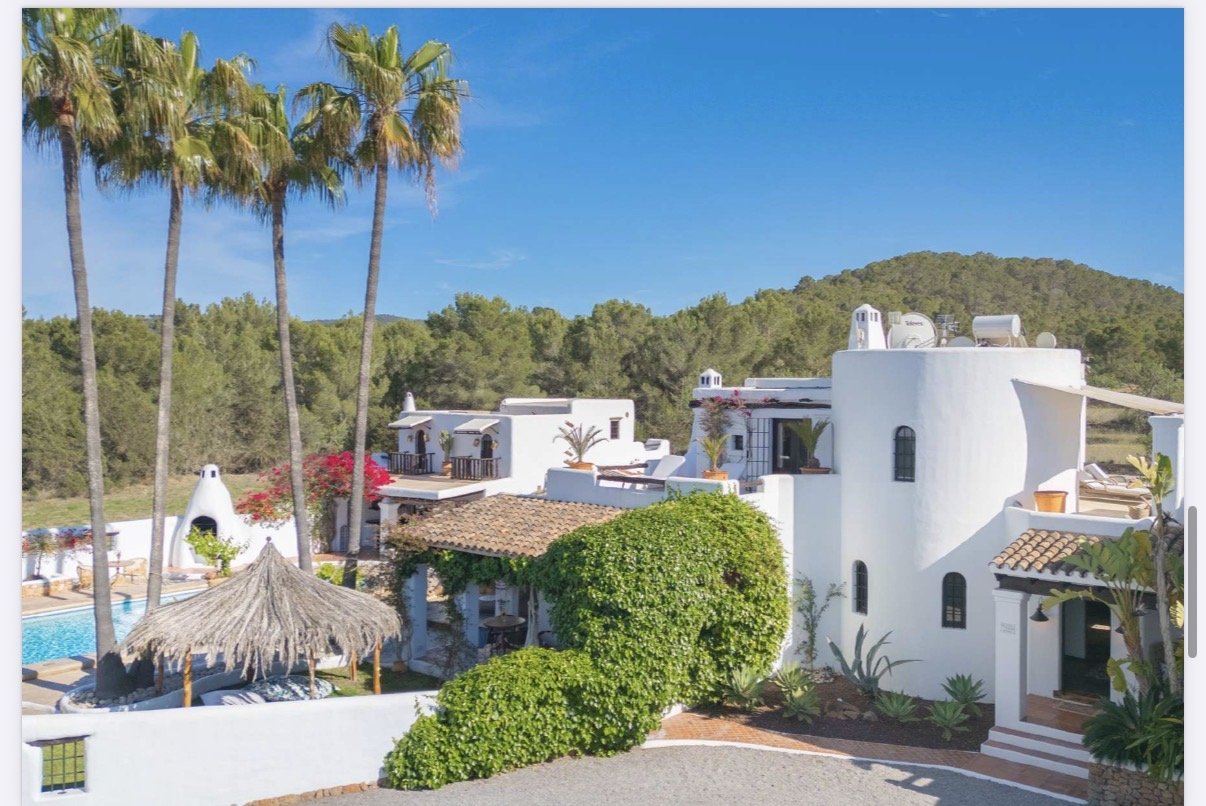 A white Mediterranean-style villa with multiple levels, curved walls, and arched windows, surrounded by green bushes and tall palm trees, with a swimming pool and thatched-roof lounging area in the yard, under a clear blue sky.