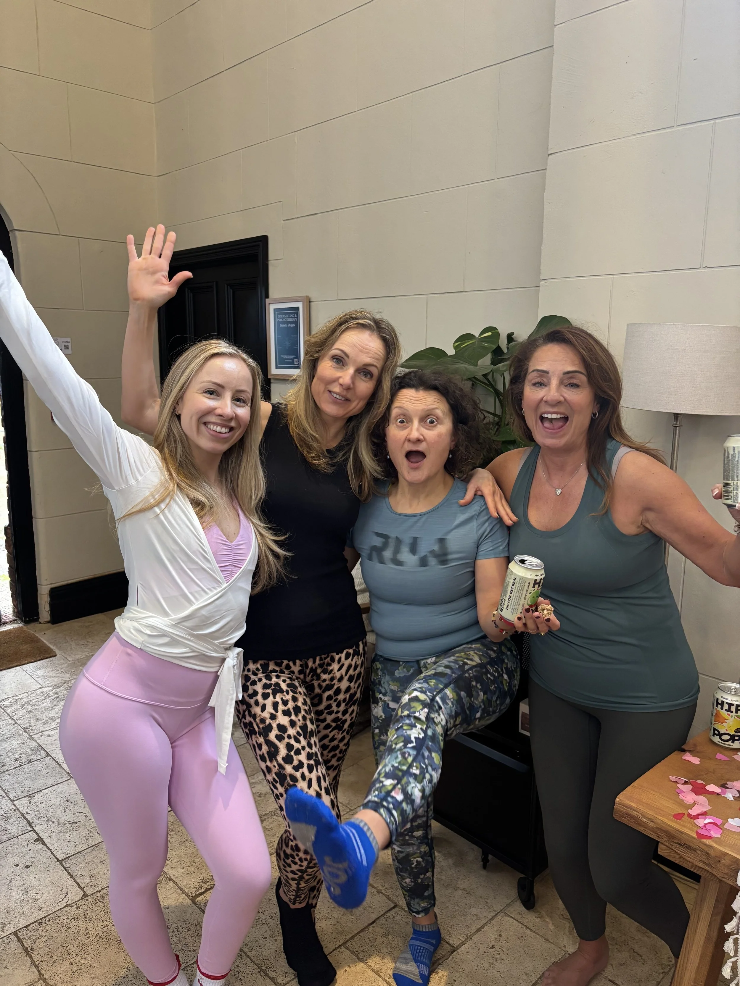 Four women posing and celebrating indoors. One woman has her arm raised, another is holding a can of drink, and they are all smiling or surprised. There are some pink paper hearts on a table in the corner.