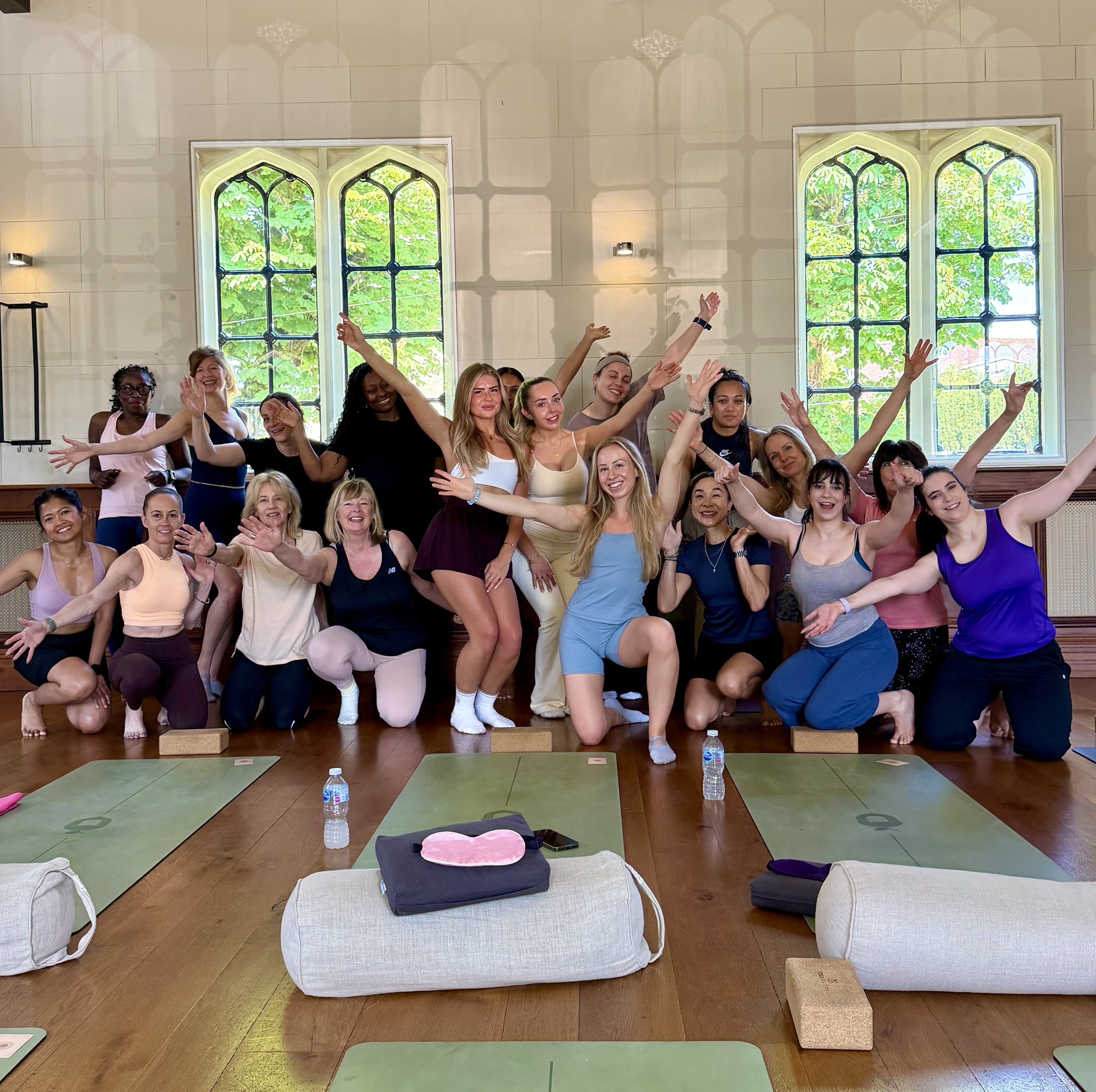 A large group of women of various ages and ethnicities posing together indoors, smiling and raising their hands during a yoga or fitness class, with yoga mats and water bottles on the floor in front and large windows with greenery outside in the background.