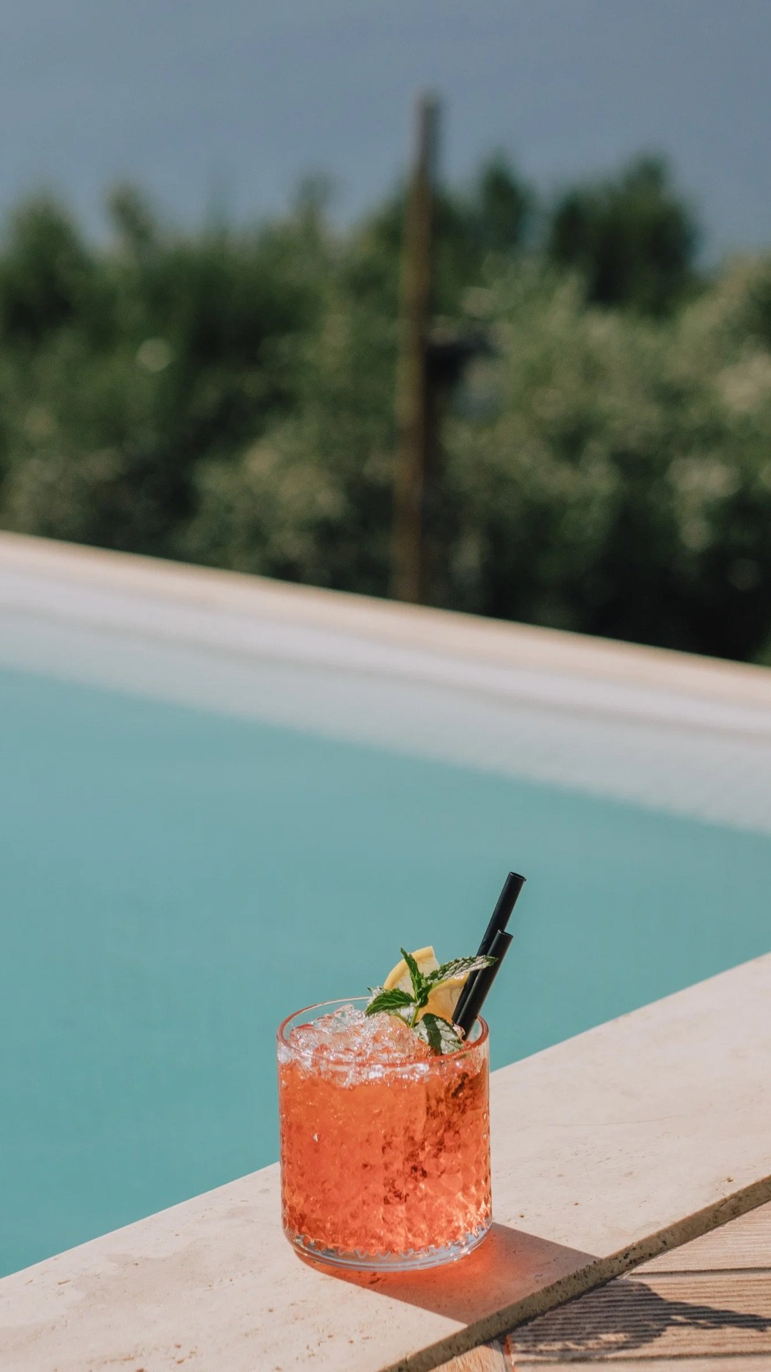 A pink cocktail garnished with a lemon wedge, mint leaves, and two black straws on a white surface near a swimming pool with a background of trees and sky.