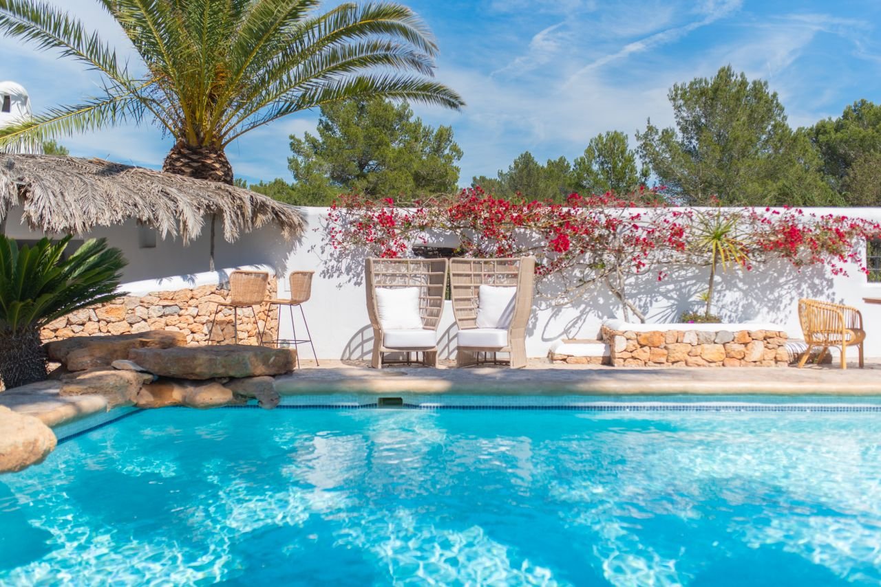 Relaxing backyard pool area with white lounge chairs, a thatched umbrella, palm trees, and flowering bushes under a partly cloudy blue sky.