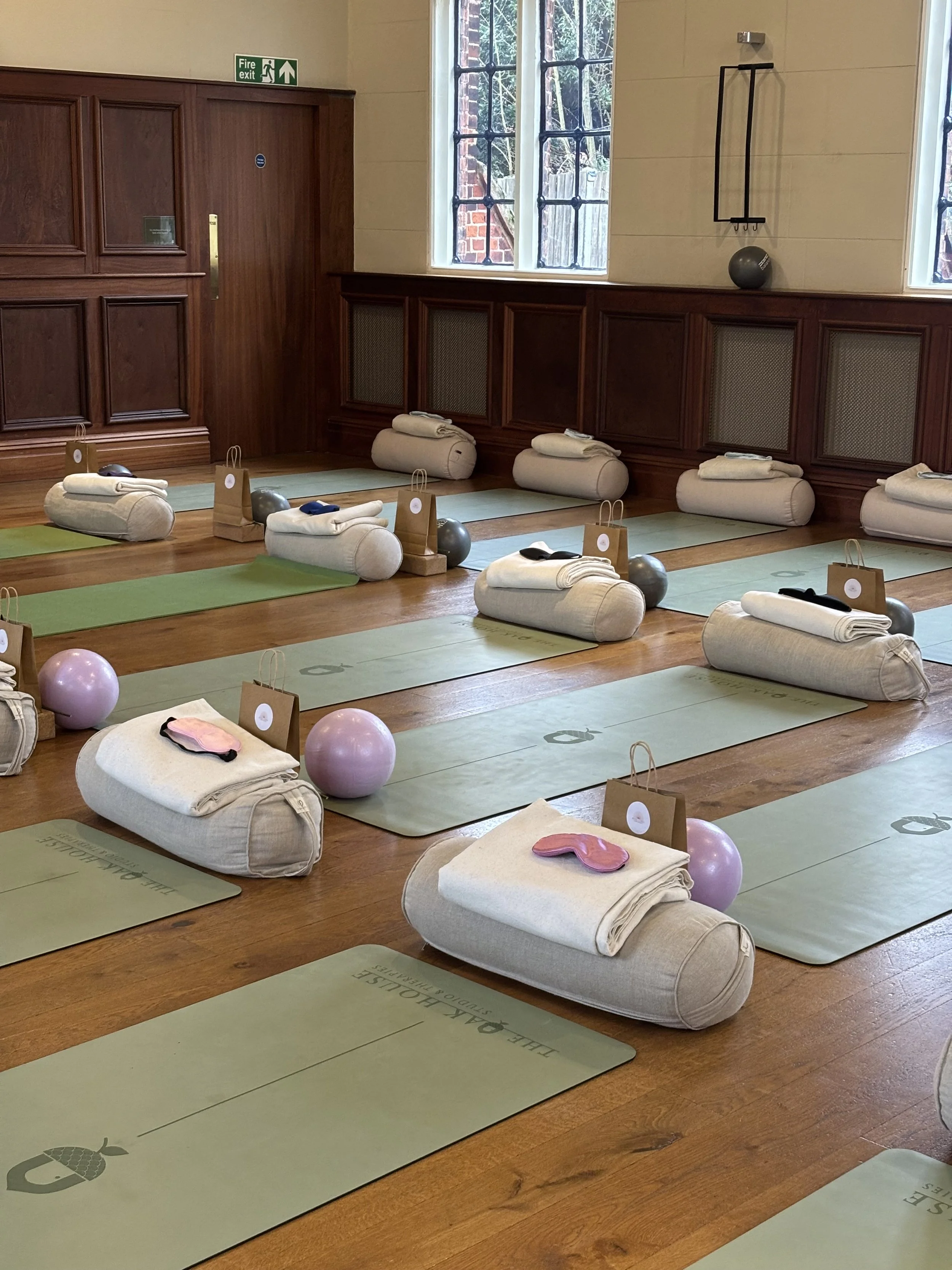 Yoga mats with rolled towels, eye pillows, small gift bags, and exercise balls arranged on a wooden floor in a room with large windows and wooden wainscoting.