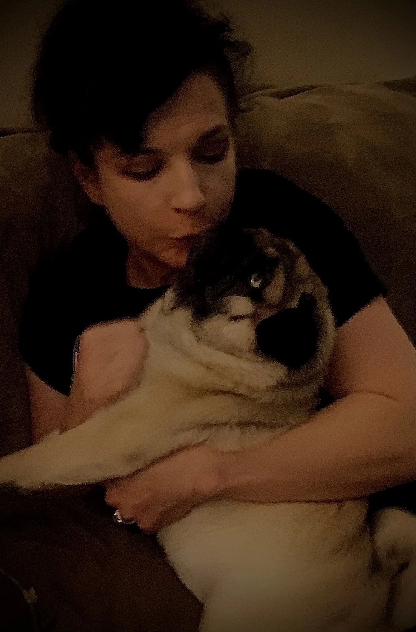 A young person with short dark hair cuddling a large dog on a brown couch in a dimly lit room.