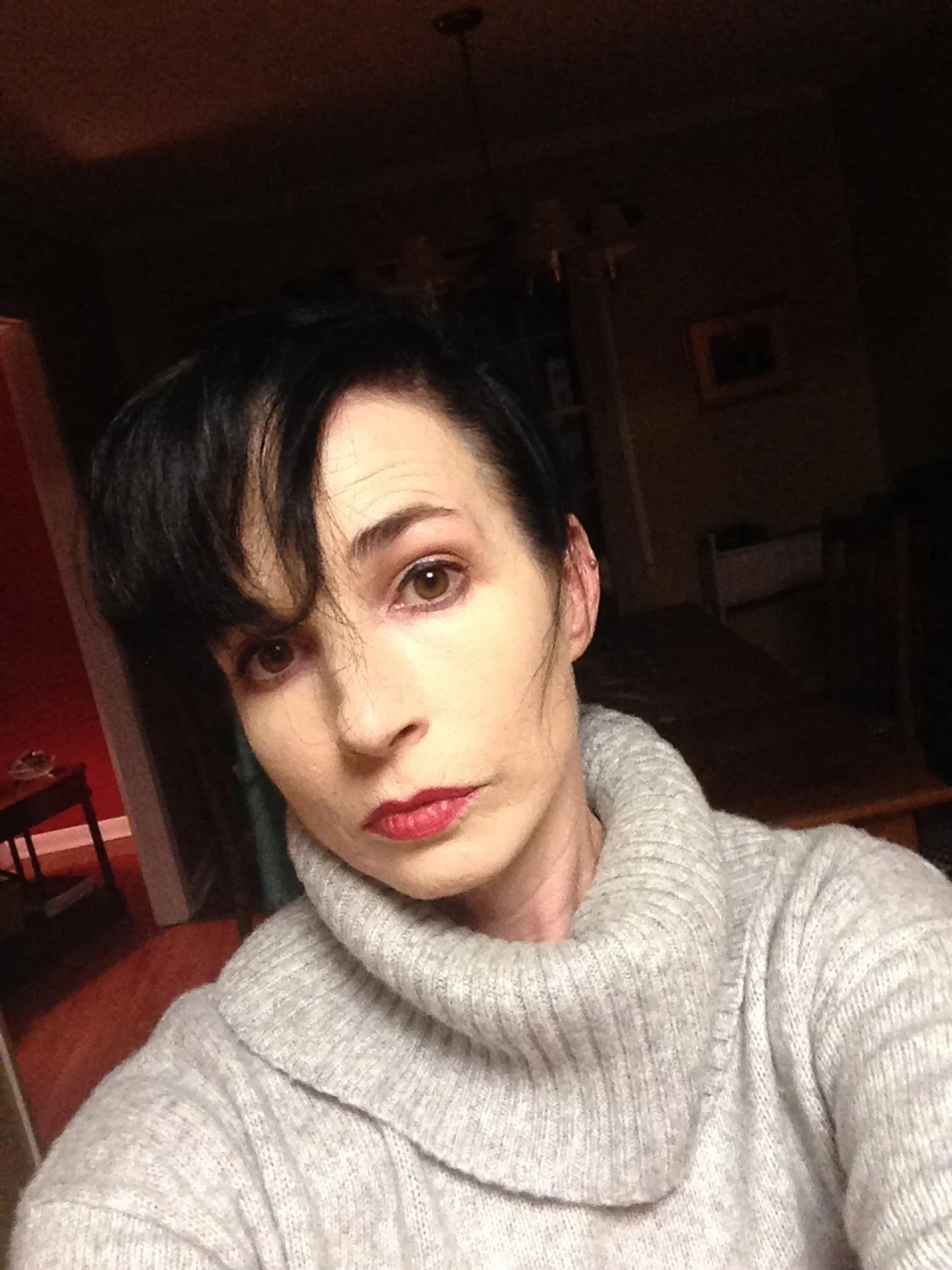 A woman with short dark hair and red lipstick wearing a light gray turtleneck sweater taking a selfie in a dimly lit room.