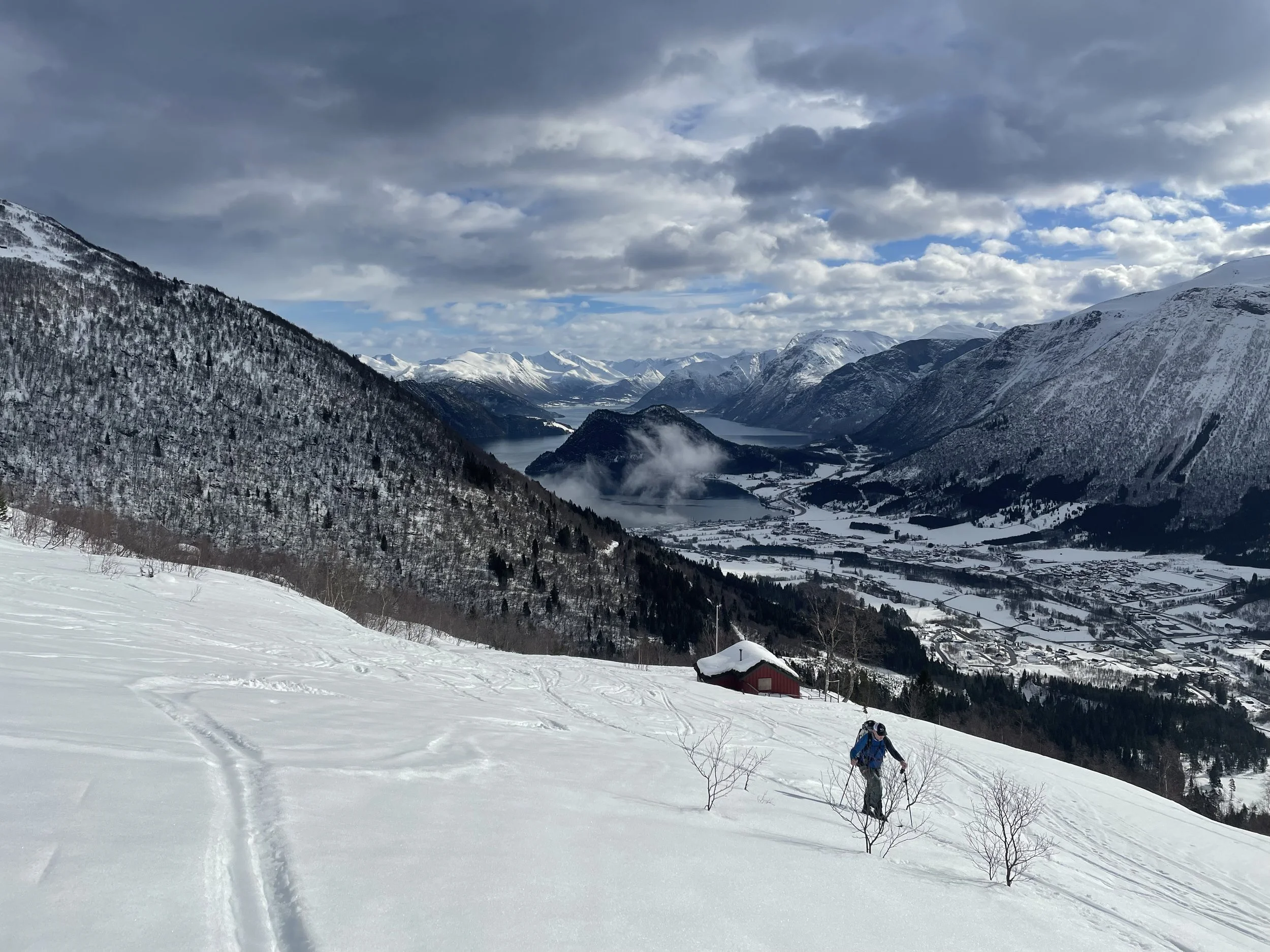 A person hiking through snow on a mountain slope with a snow-covered cabin nearby. In the background, there are snow-capped mountains, a lake, and a valley with scattered houses under a cloudy sky.